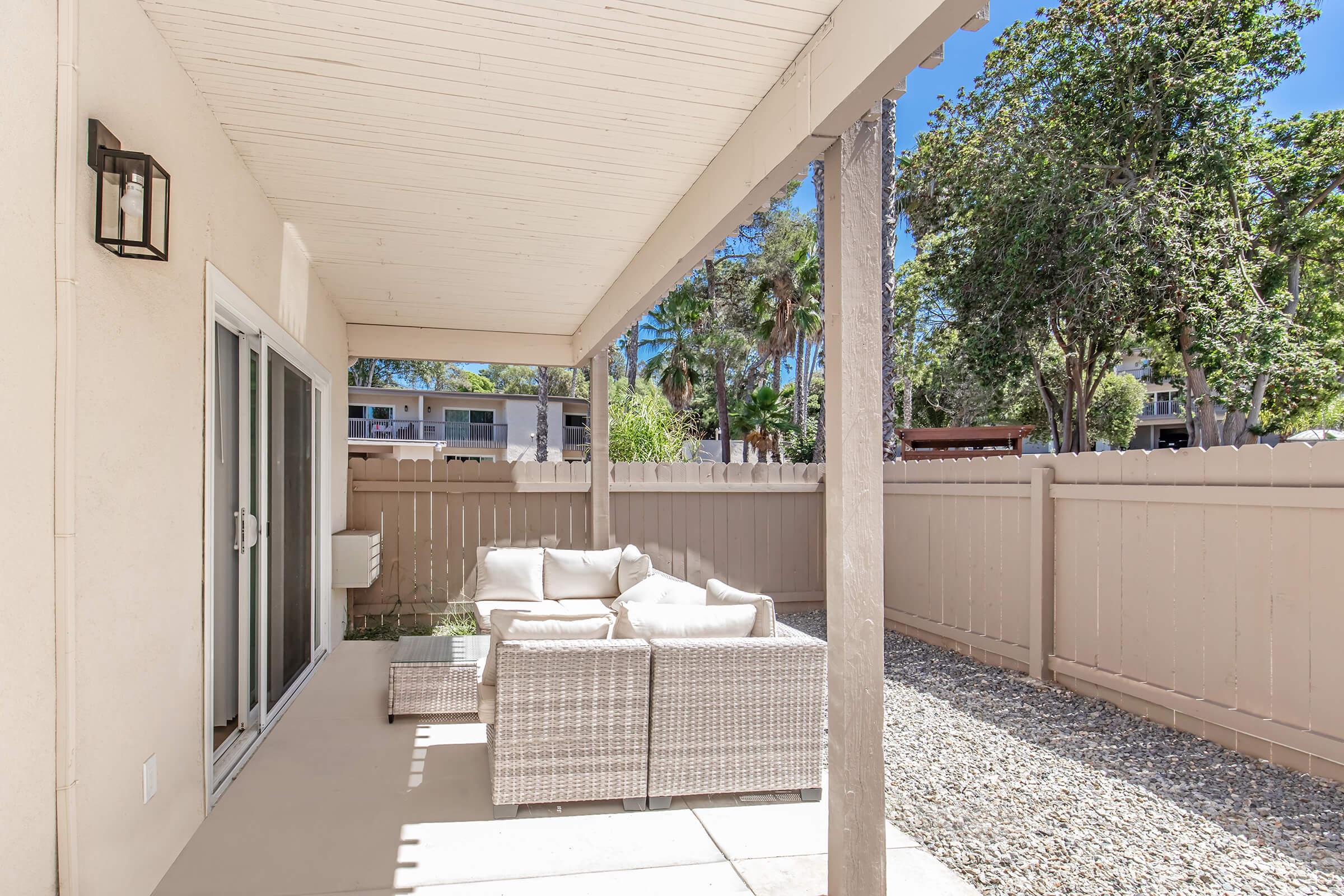 A cozy outdoor patio area featuring a covered seating arrangement with white cushions and a small coffee table. The space is surrounded by a beige fence and lush greenery, providing a tranquil atmosphere. Sunlight streams in, highlighting the inviting area ideal for relaxation or social gatherings.