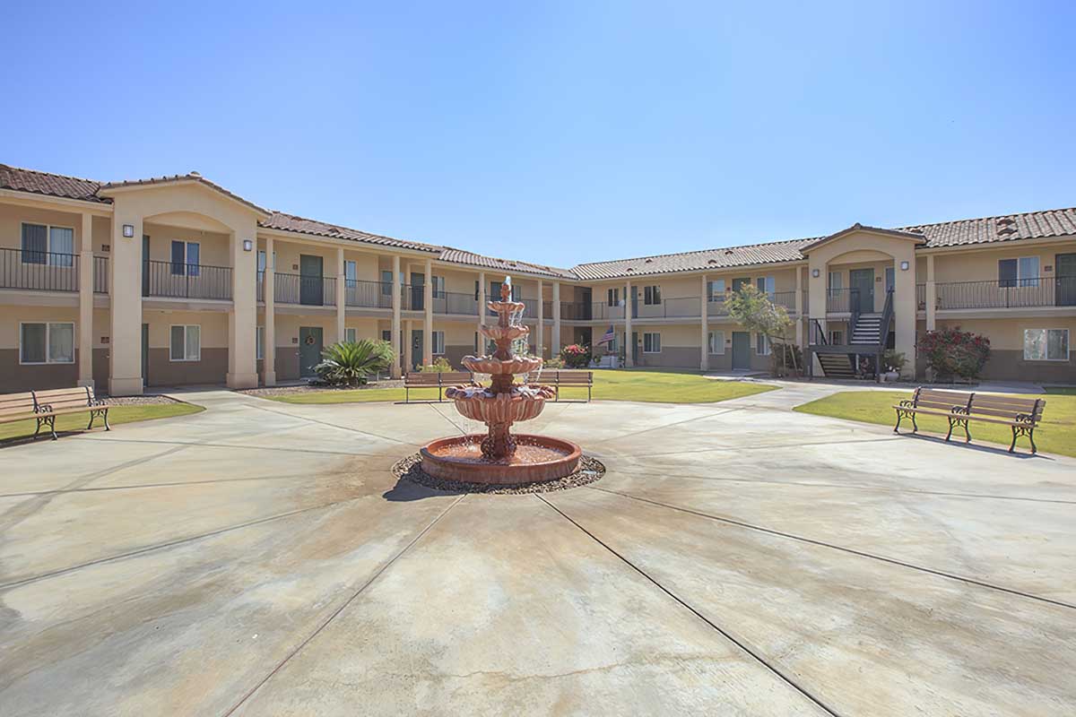 A central fountain is surrounded by a landscaped courtyard with benches and greenery, flanked by two-story apartment buildings.