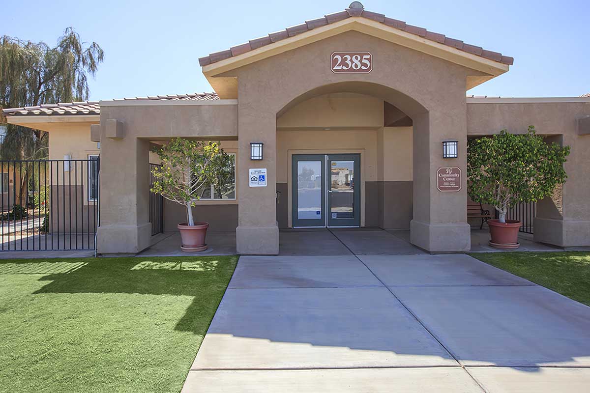 Front view of a single-story building with a large entrance featuring double doors. The structure has a light brown exterior with decorative archways, two large planters with greenery, and a smooth concrete walkway leading to the entrance.