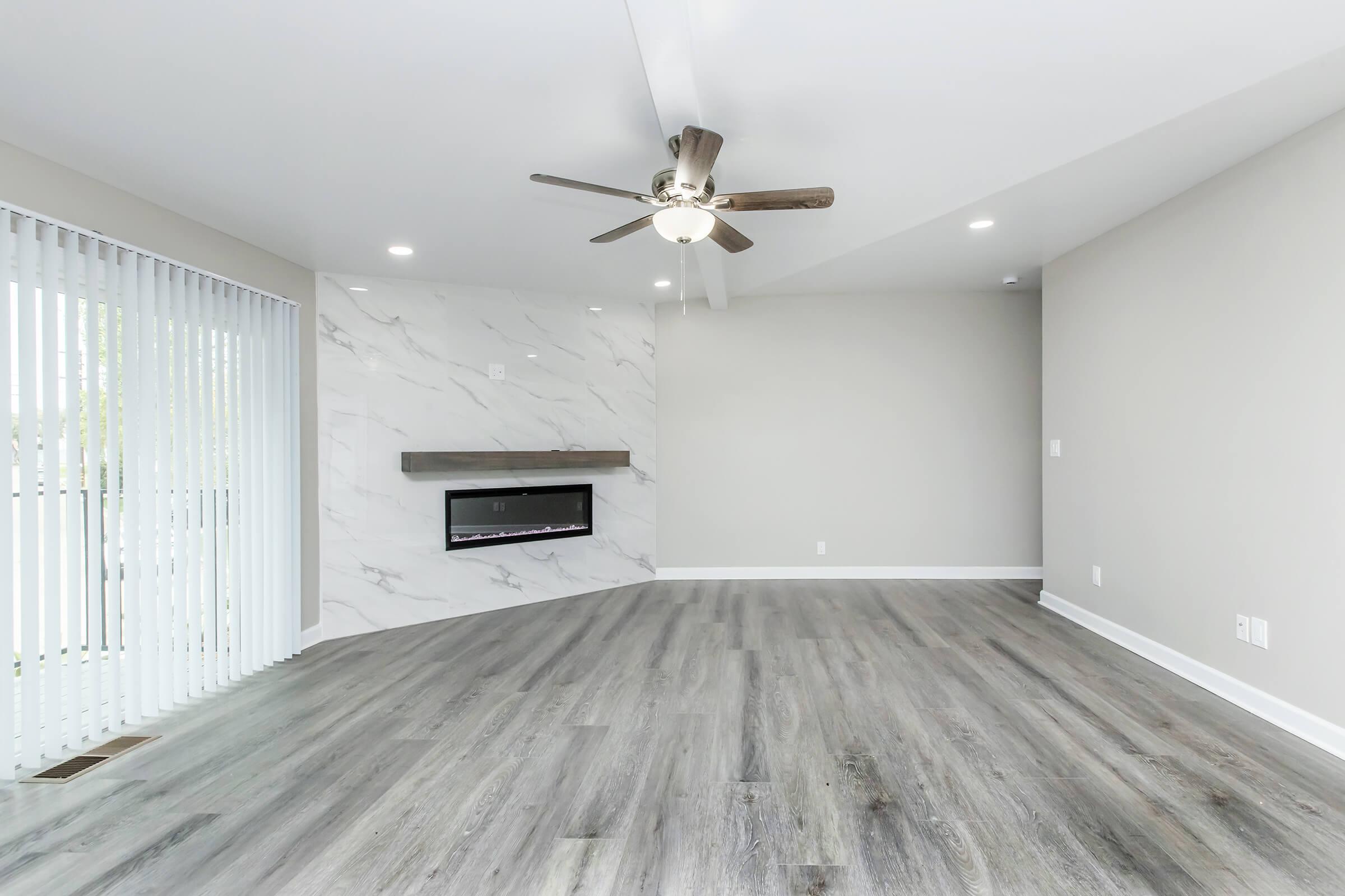 A modern living room featuring a sleek ceiling fan, an elegant wall-mounted fireplace trimmed in marble, and large sliding glass doors with vertical blinds. The room has light gray walls and a spacious, open layout with durable wood-like flooring. Natural light floods in from the sliding doors, creating a bright atmosphere.