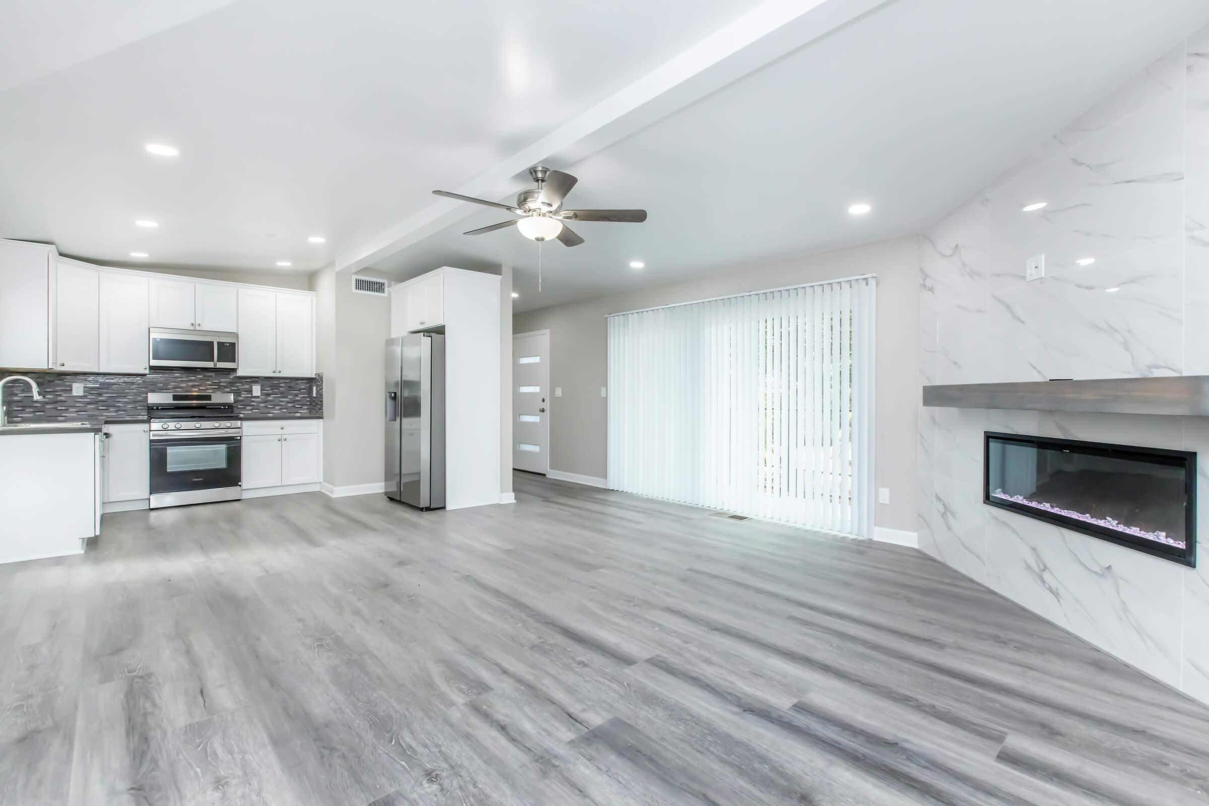 A modern, spacious living area featuring an open kitchen with white cabinets, stainless steel appliances, and a sleek design. The room has grey wood-like flooring, a ceiling fan, and large windows with vertical blinds, along with a contemporary fireplace on one wall.