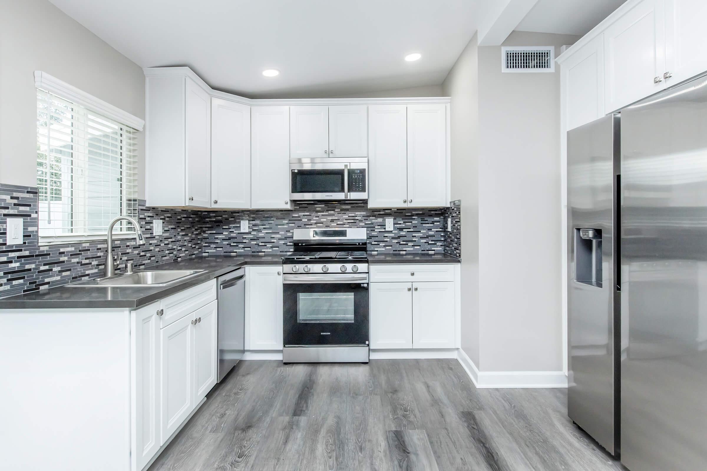 Modern kitchen featuring white cabinetry, stainless steel appliances, a sleek black and gray tiled backsplash, and hardwood-style flooring. Natural light floods the space through windows, enhancing the contemporary design.