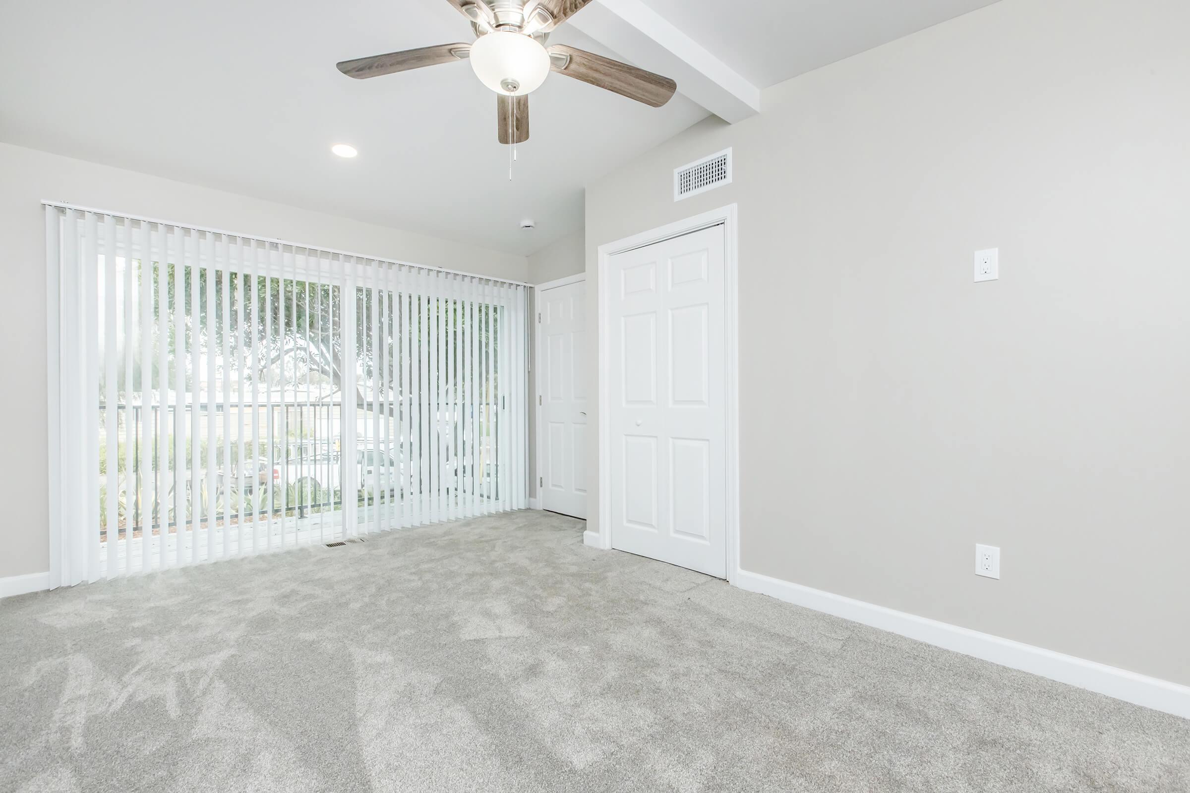 A well-lit interior room with light gray carpet, a ceiling fan, and white vertical blinds covering large windows. A white closet door is visible on the right, and walls are painted in a soft gray color. The space appears clean and inviting, suitable for various uses.