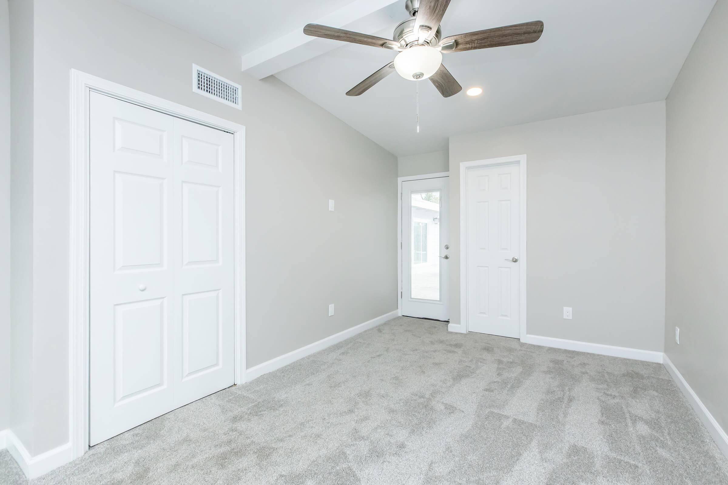A well-lit, empty room featuring light gray walls and plush beige carpet. The room has a ceiling fan with wooden blades, a closet with double doors, and a door leading outside. Natural light enters through the door, creating a bright and inviting space.