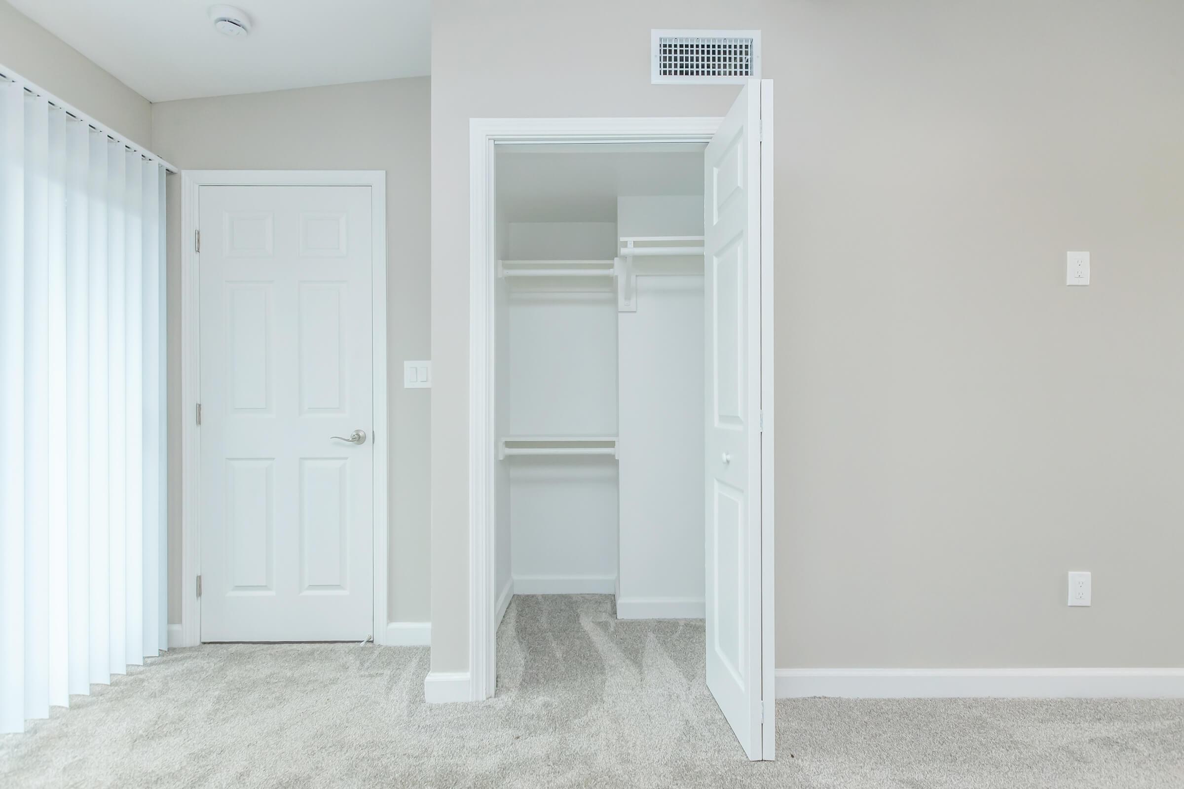 A modern, minimalistic room featuring light gray walls and a carpeted floor. The space includes a closed white door and an open closet with shelves, showcasing a bright and airy atmosphere enhanced by natural light from the window.