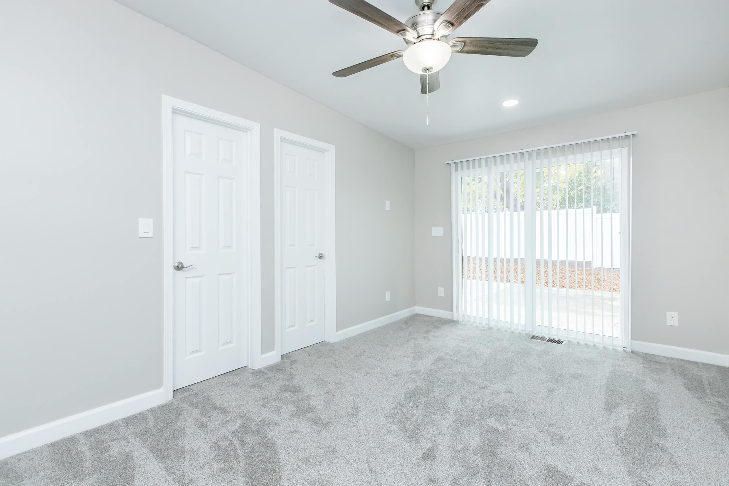 Interior view of a freshly painted room featuring light gray walls and plush carpet. It includes two white doors on the left, a ceiling fan with light fixture in the center, and a large window with vertical blinds allowing natural light. The view outside shows a landscaped area.