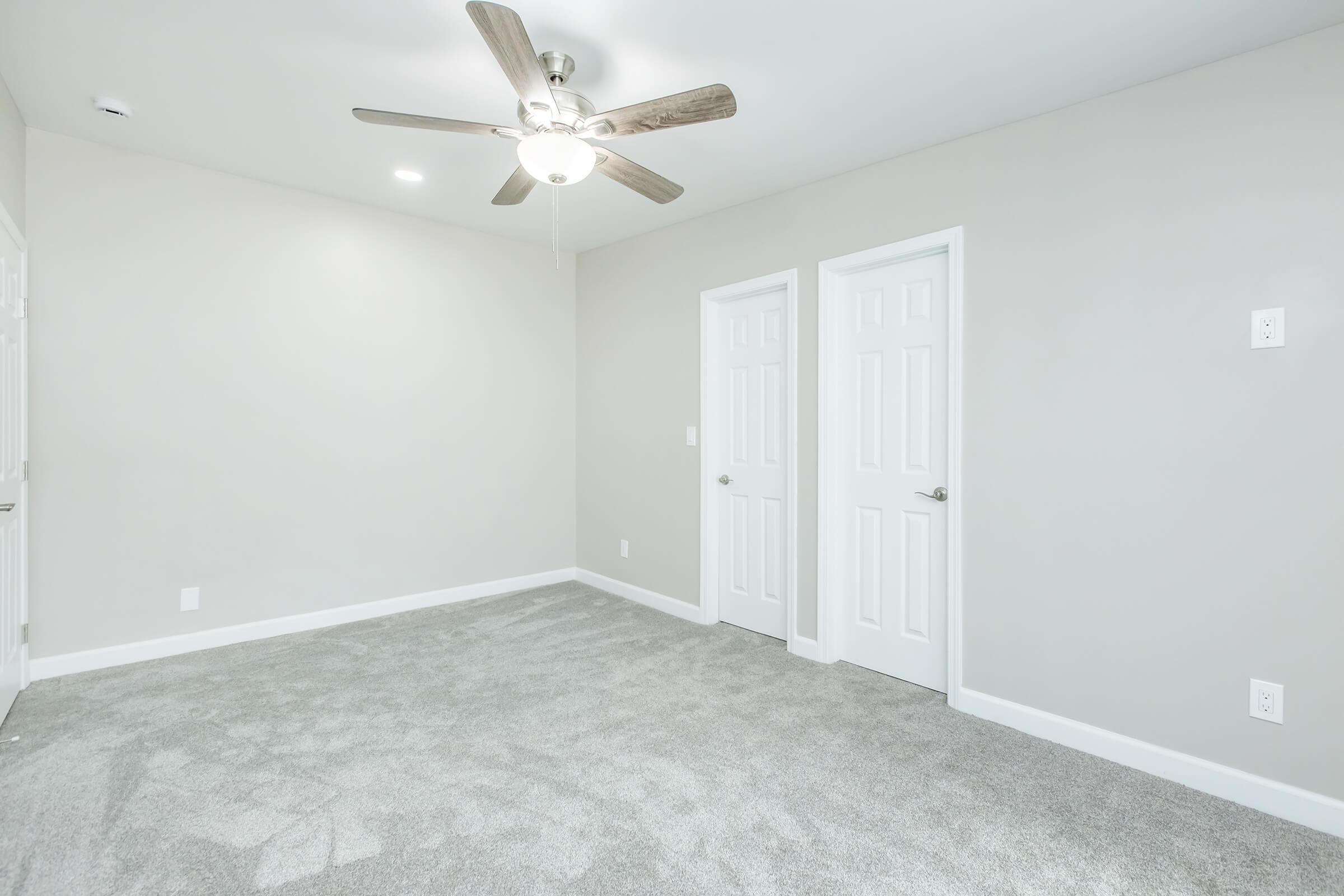 Empty room featuring light gray walls, a ceiling fan with wooden blades, and plush gray carpeting. Two closed white doors are on the right side, with a small white wall switch visible. The room is well-lit and uncluttered, creating a clean and simple aesthetic.