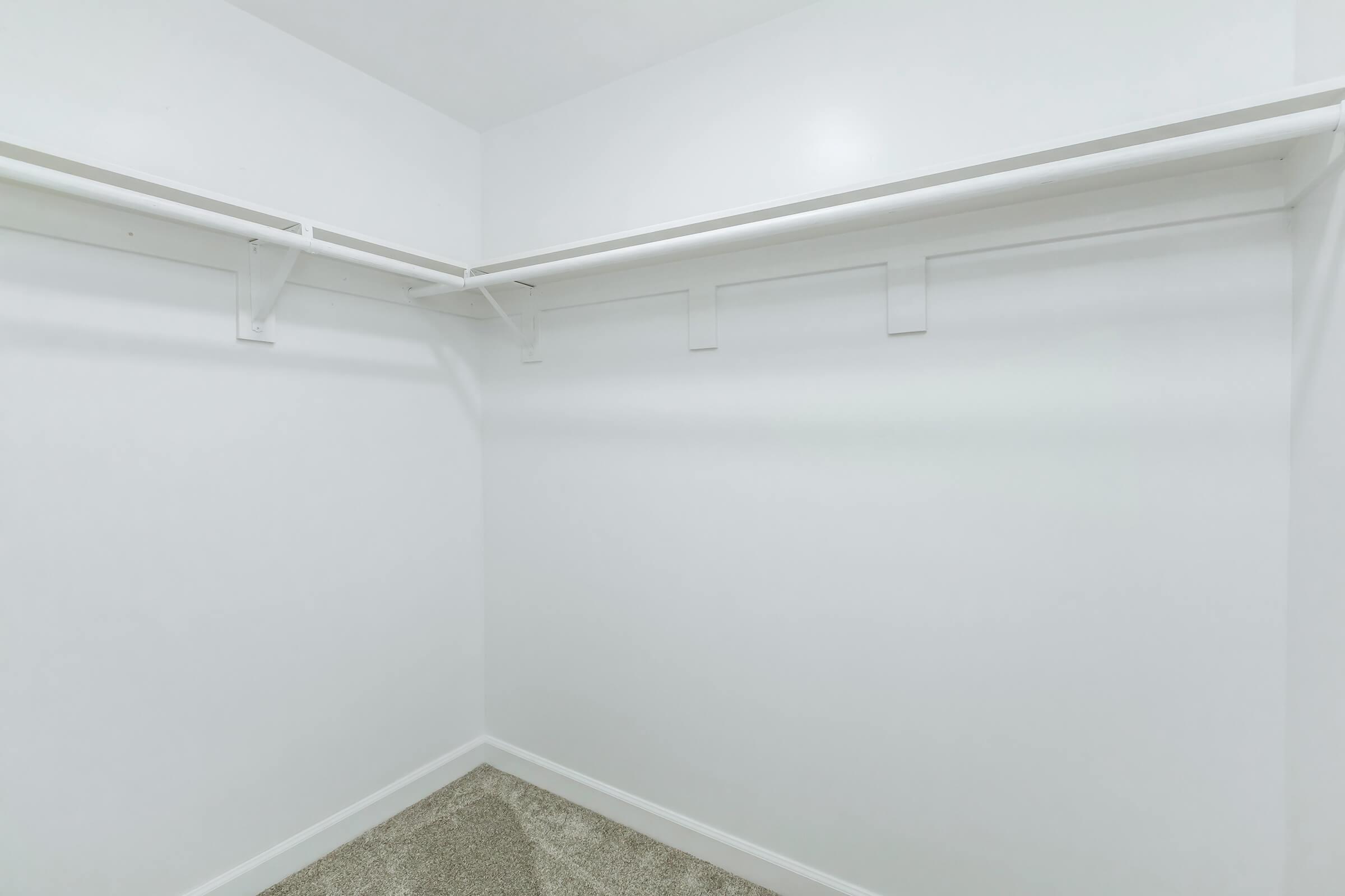Empty closet with white walls, a light-colored carpeted floor, and a simple white shelf and hanging rod. The space is well-lit and uncluttered, offering ample room for storage or organization.