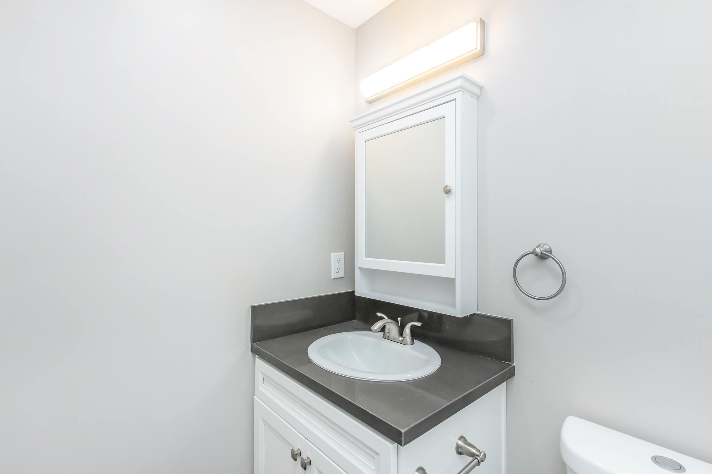 A modern bathroom featuring a white vanity with a sink, a mirrored cabinet above, a gray countertop, and a wall-mounted towel ring. A toilet is visible to the right, and the walls are painted a light gray. A wall light fixture provides illumination above the mirror.
