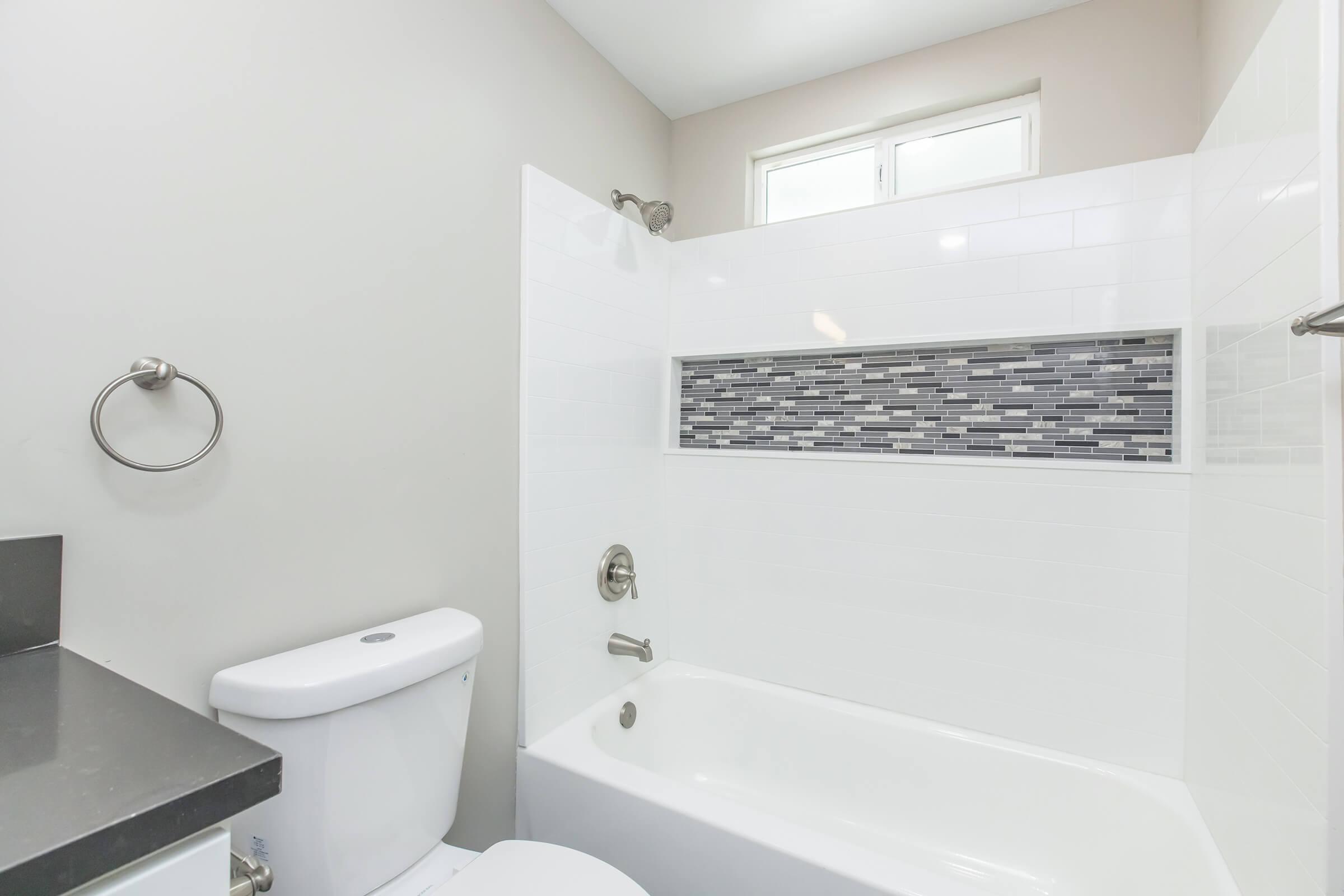 A modern bathroom featuring a white bathtub with a tiled backsplash, a chrome faucet, and a wall-mounted towel holder. The bathroom has light gray walls, a window for natural light, and a sleek countertop next to a white toilet.