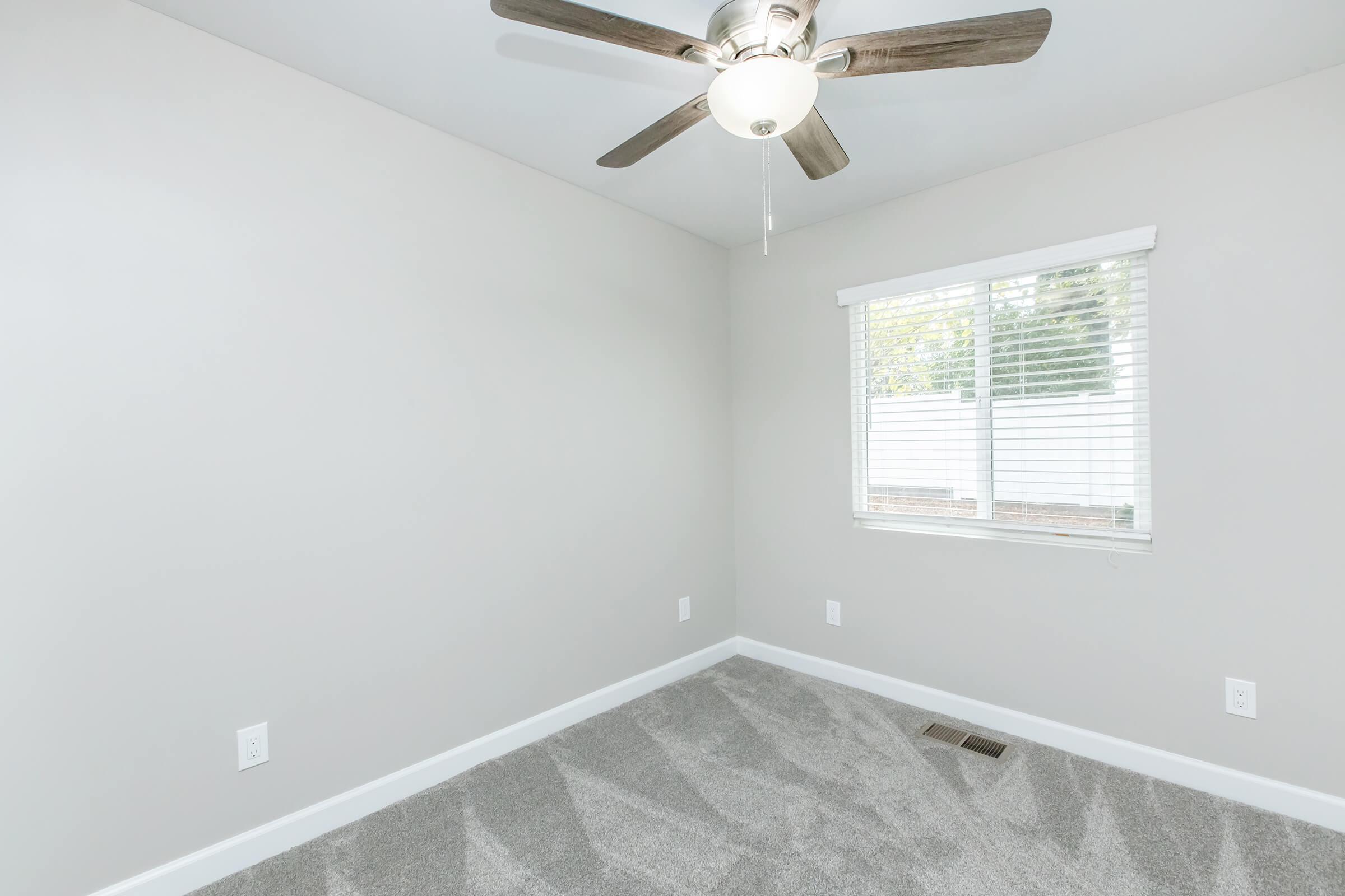 Empty room with light gray walls and carpet. A ceiling fan with wooden blades hangs from the ceiling. A window with white blinds allows natural light in, and a small vent is visible on the floor. The room is unfurnished, creating a spacious and clean atmosphere.