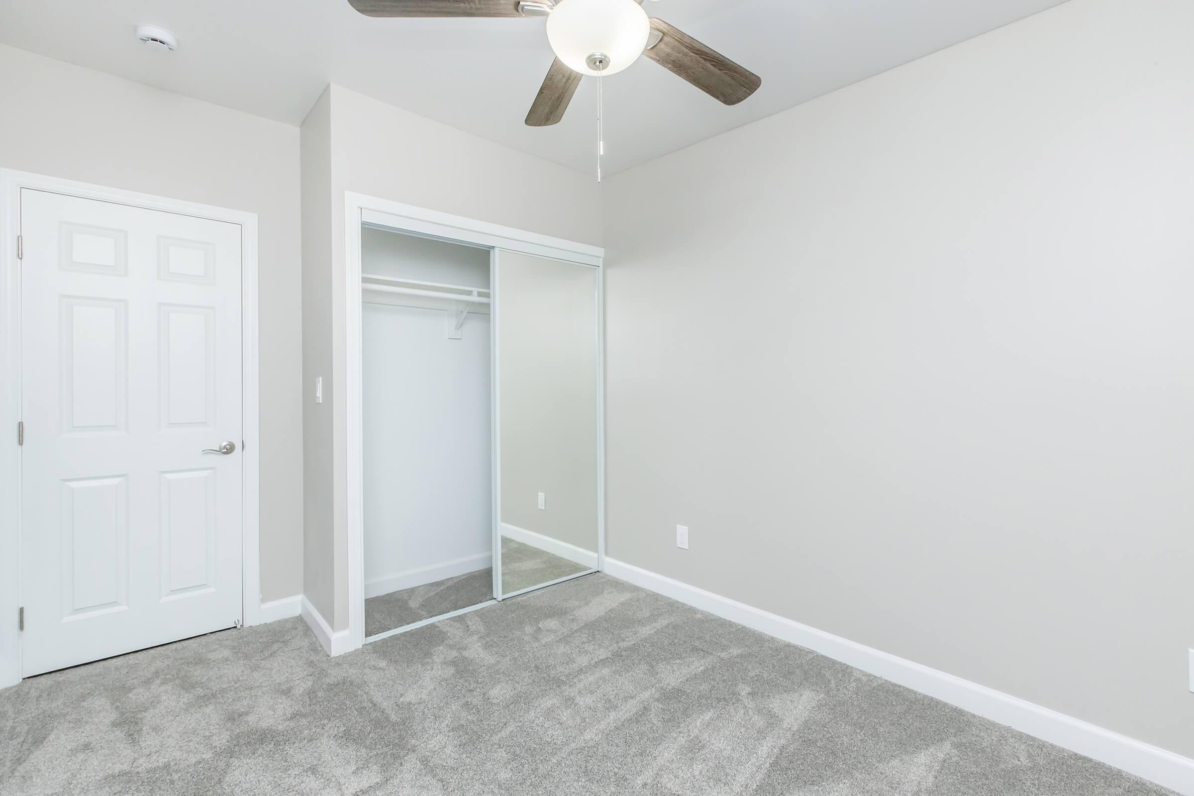 A minimalist room featuring light gray walls and plush carpet. A white closet with mirrored sliding doors is visible on the left. The room has a ceiling fan with wooden blades and a simple light fixture. A closed white door can be seen in the corner, adding to the room's clean and spacious appearance.