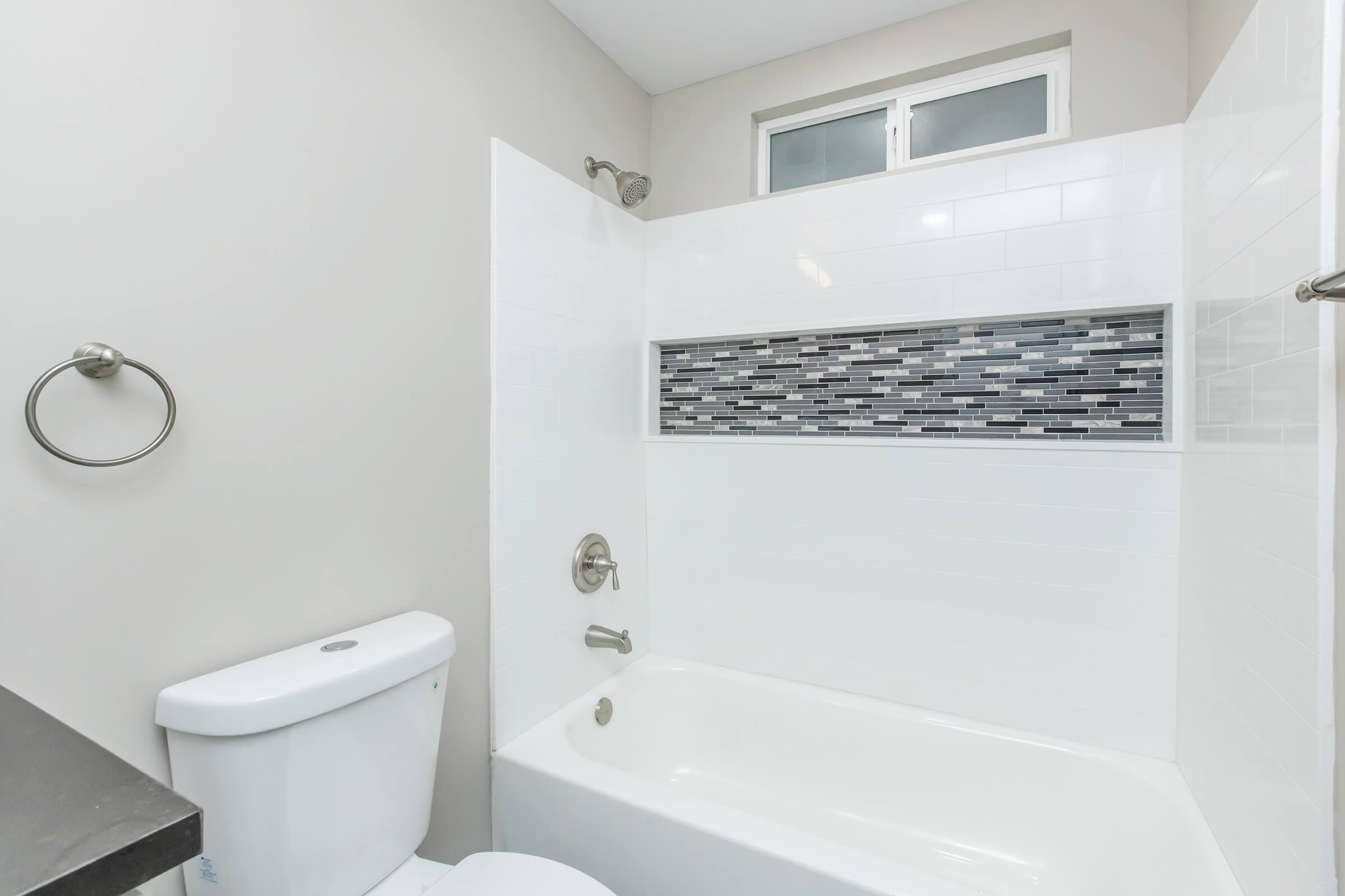 A clean, modern bathroom featuring a white bathtub with a tiled accent wall in shades of gray and black. The space includes a toilet, a sleek faucet, and a towel ring mounted on the wall. Natural light shines through a window above the tub, enhancing the airy feel of the room.