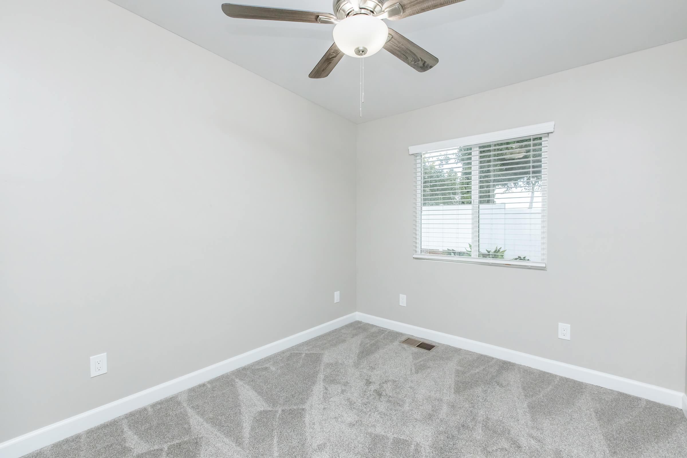 A minimalistic empty room with light gray walls and soft carpet. There is a white window with blinds letting in natural light, and a ceiling fan with wooden blades. The room features baseboards and electrical outlets, creating a clean and modern aesthetic.