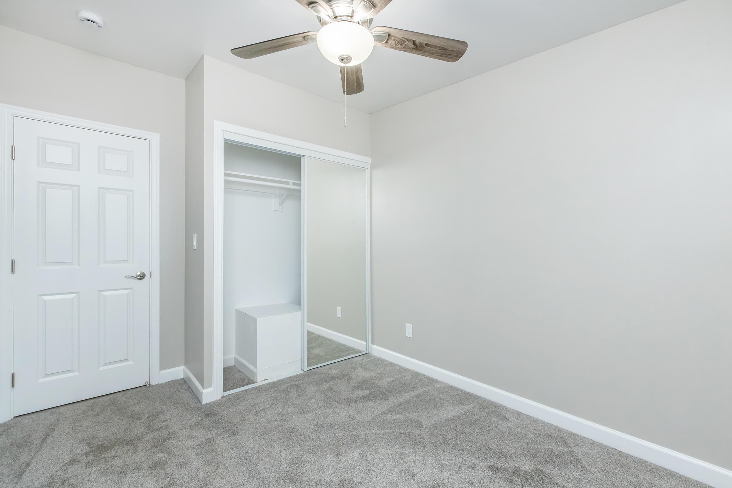 A minimalist room featuring light gray walls and new carpet, with a ceiling fan and a closed white door. There is an open closet with a white storage unit inside and a mirror reflecting part of the room. The space is well-lit and appears to be freshly painted and decorated.