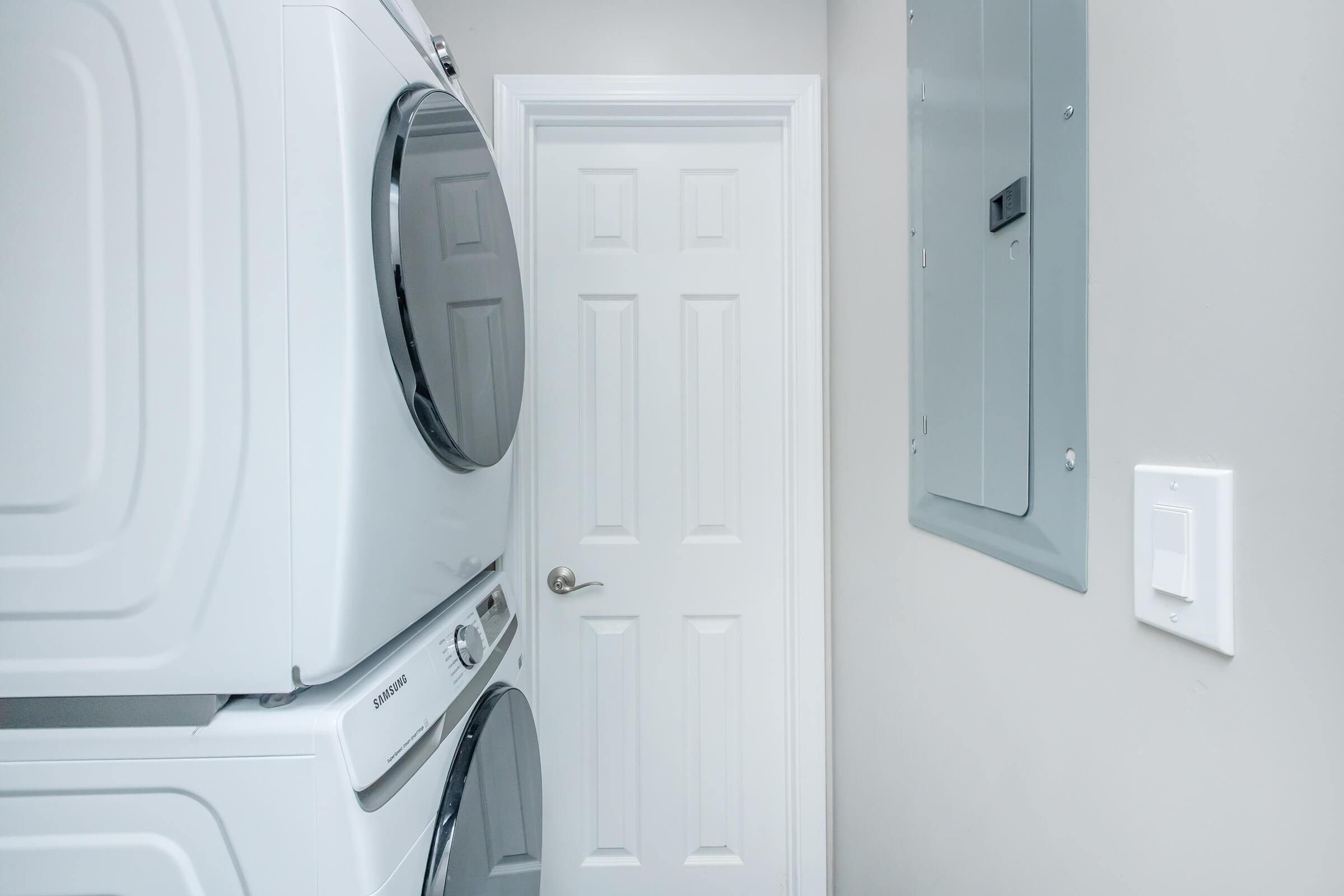 A laundry room featuring stacked white washer and dryer appliances on the left, a closed white door at the end of the hallway, and an electrical panel mounted on the wall to the right. The walls are painted in a light color, creating a clean and modern look.