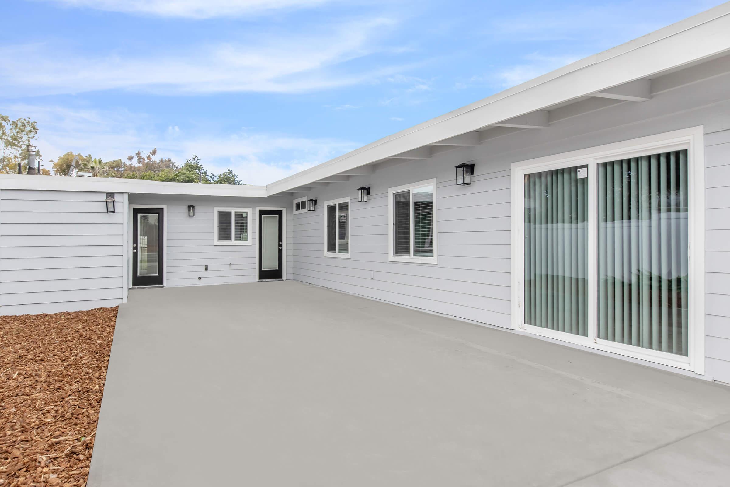A modern gray house exterior with a spacious patio area. The house features multiple doors and large windows with blinds. The surrounding area has mulch landscaping, and the sky is bright with a clear blue backdrop.