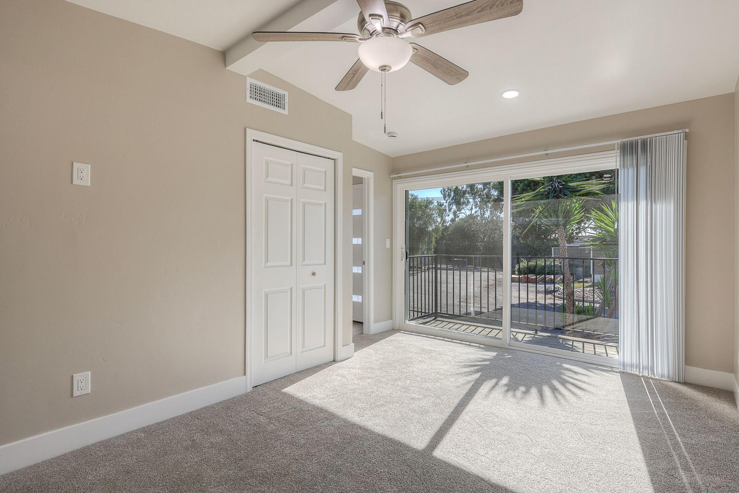 Bright interior of a room featuring light-colored walls, a ceiling fan, and large windows with views of greenery outside. The floor is carpeted, and sliding glass doors lead to a balcony. The space is well-lit, creating an inviting atmosphere.