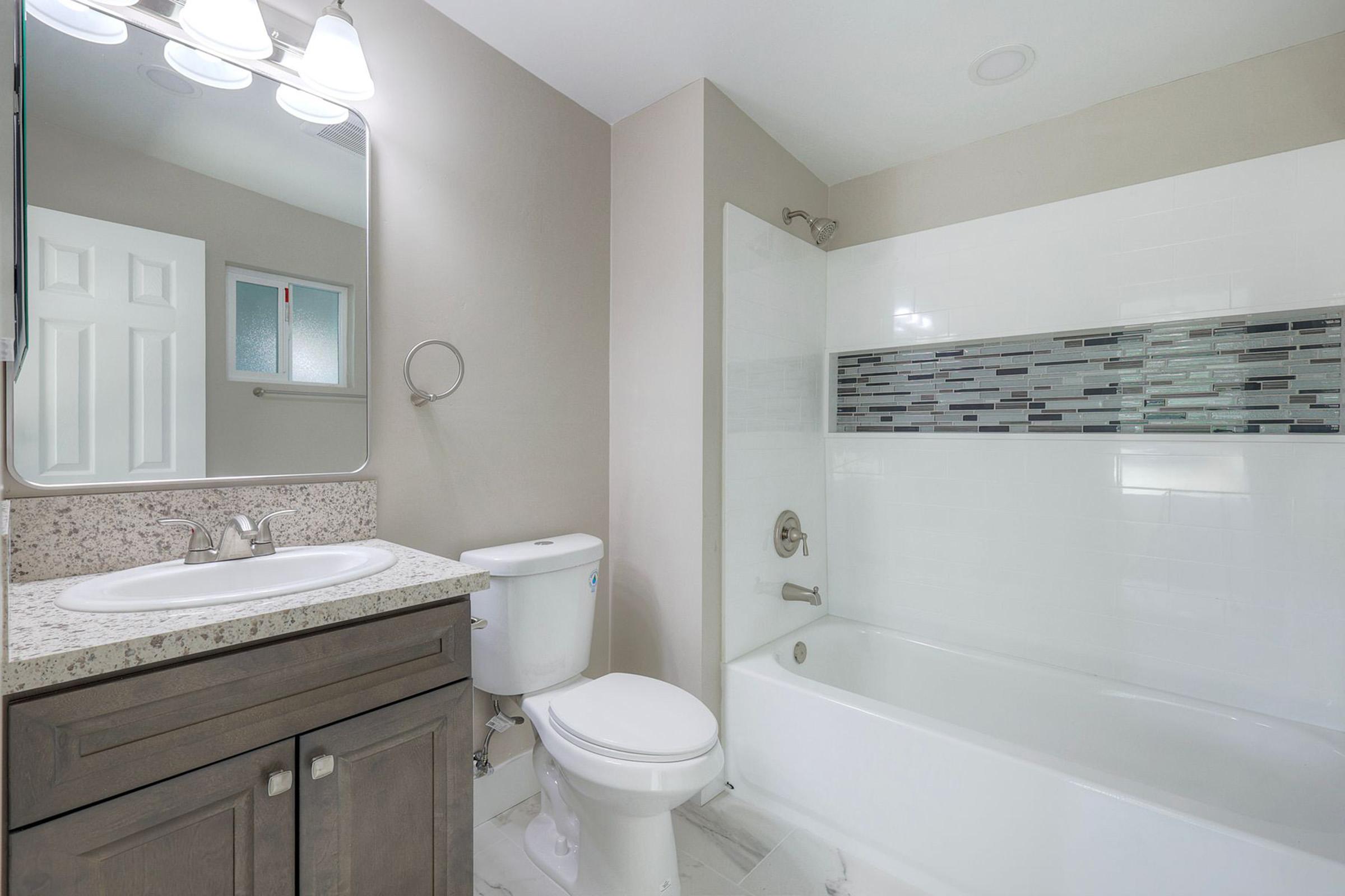 A modern bathroom featuring a white bathtub with a glass tile accent, a dark wood vanity with a granite countertop, a round mirror above the sink, a toilet, and neutral-colored walls. Natural light is coming from a window in the background.