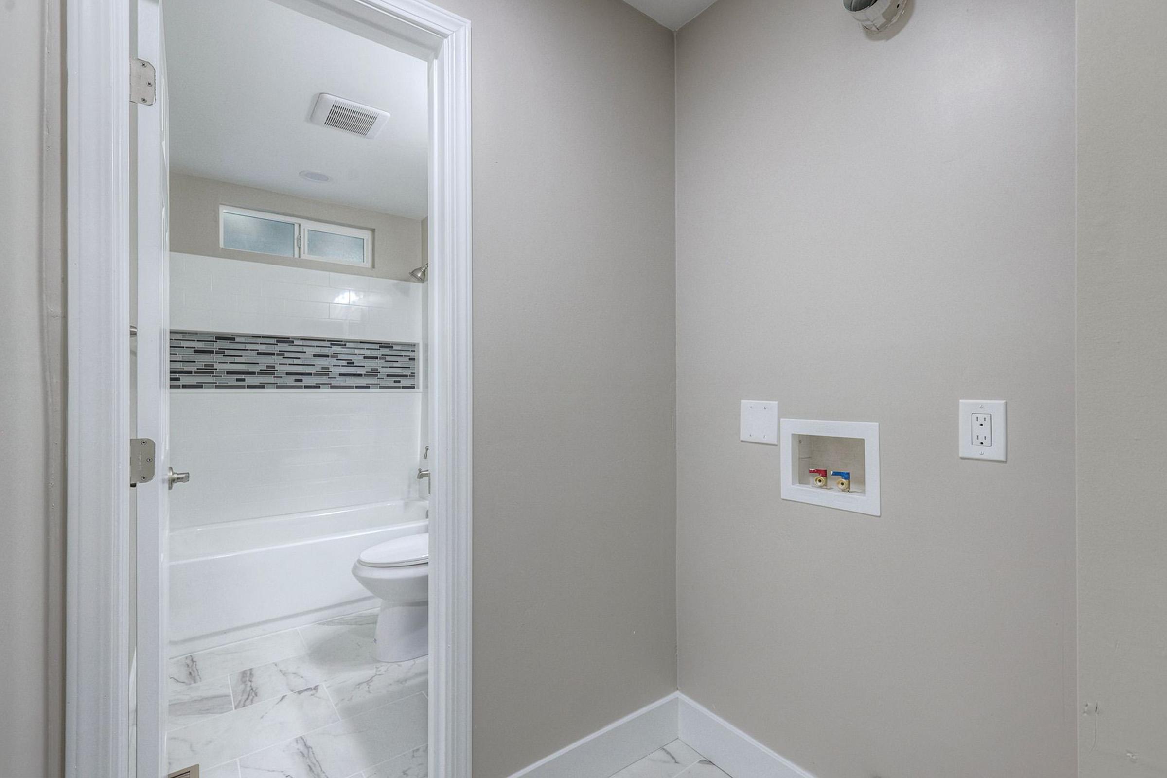 A view of a bathroom entry featuring light gray walls, a white bathtub with a tiled backsplash, and a small recessed shelf in the wall. The floor is covered with marble tiles, and there is a doorway leading to the bathroom. A light fixture is visible on the right side of the image.