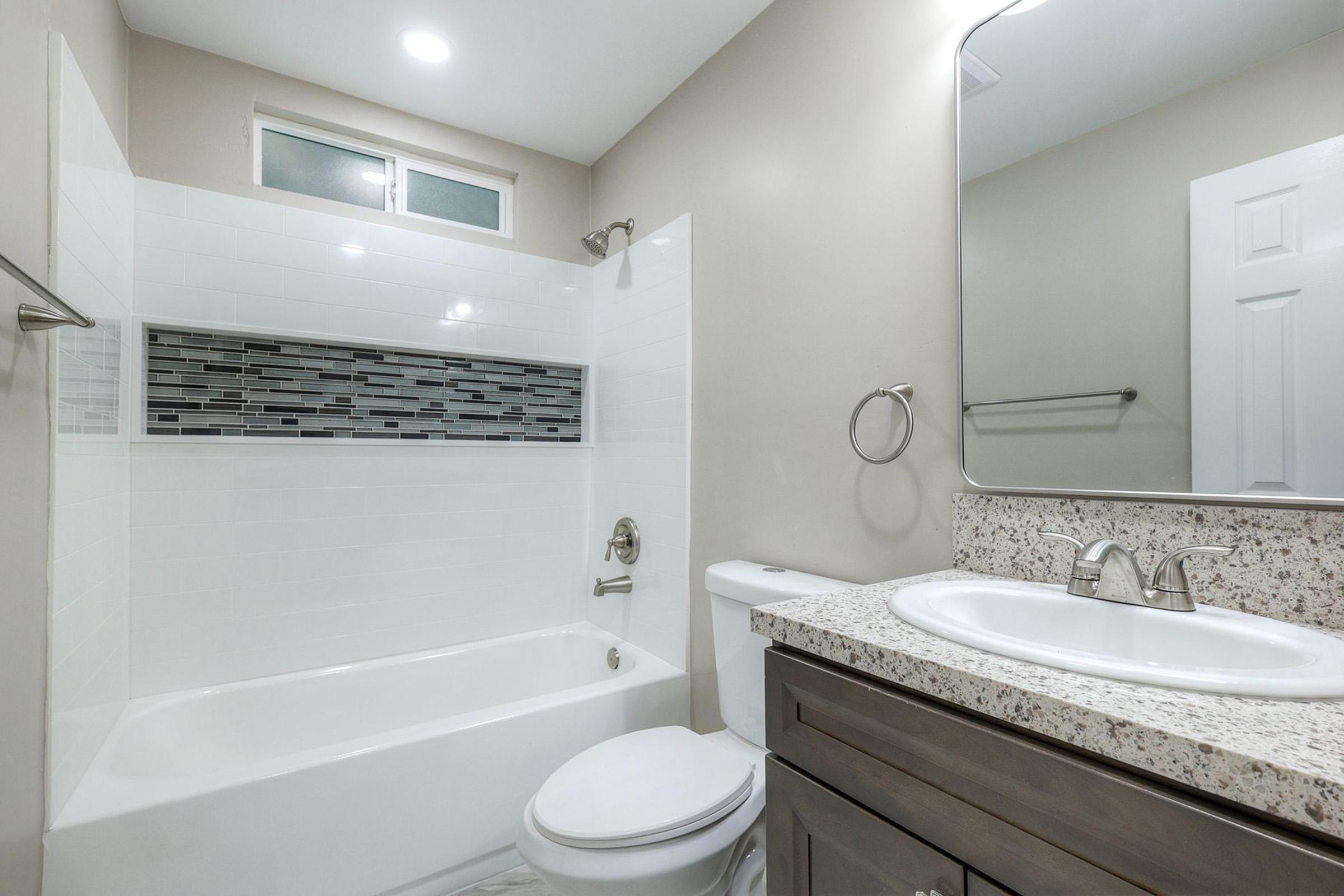 A modern bathroom featuring a bathtub with a tiled backsplash, a granite countertop sink with two faucets, a large mirror, and a toilet. The walls are painted in neutral colors, and there is natural light coming through a small window above the bathtub.