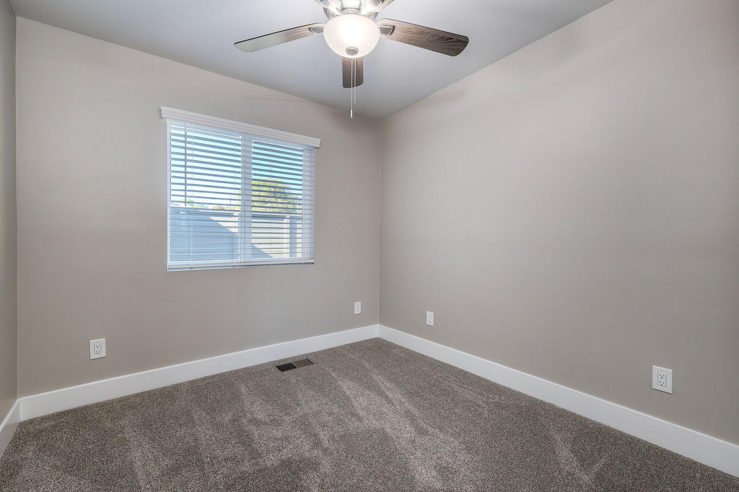 A small, empty room featuring beige walls, a ceiling fan, and a window with blinds. The floor is covered with light gray carpet, and there are no furnishings. There are two electrical outlets and a vent on the floor, providing a simple and clean aesthetic.