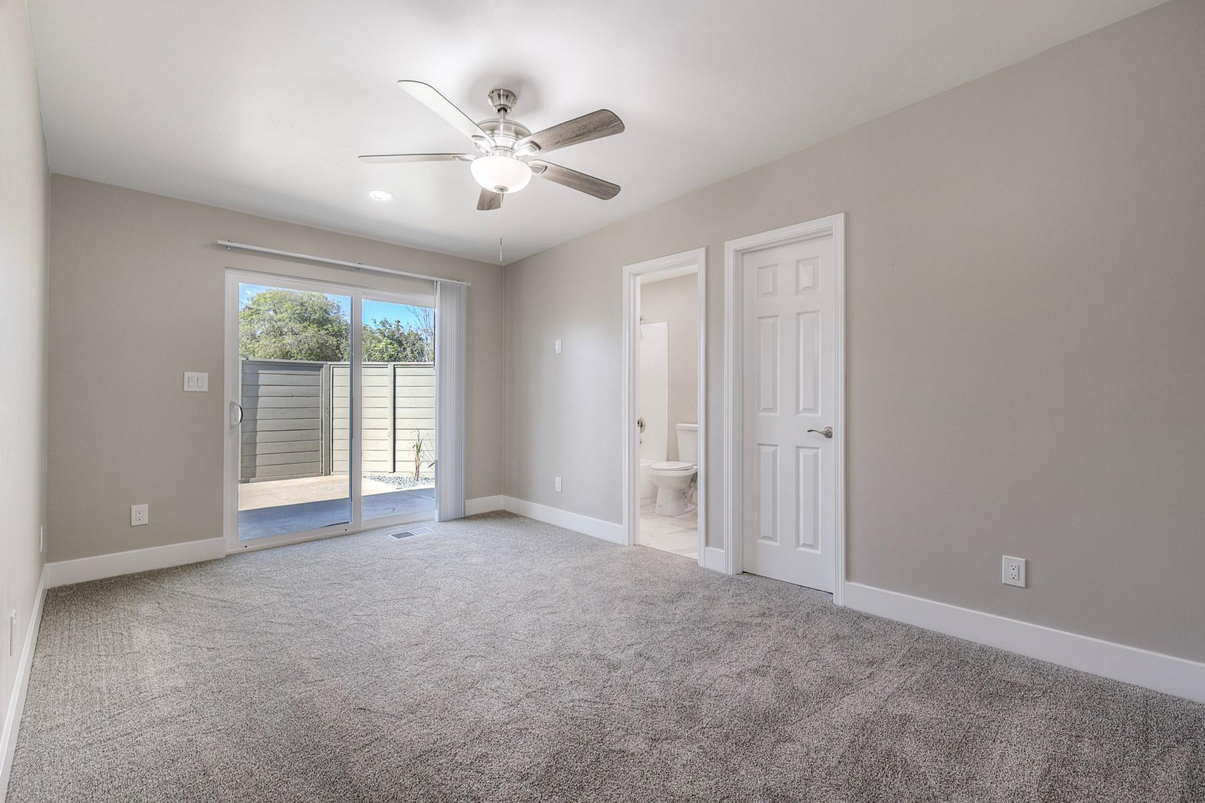 Spacious, light-filled room with a ceiling fan, neutral gray walls, and plush carpeting. Features a large sliding glass door leading to an outdoor area, a doorway to a bathroom, and a closed door, creating a cozy and modern living space.