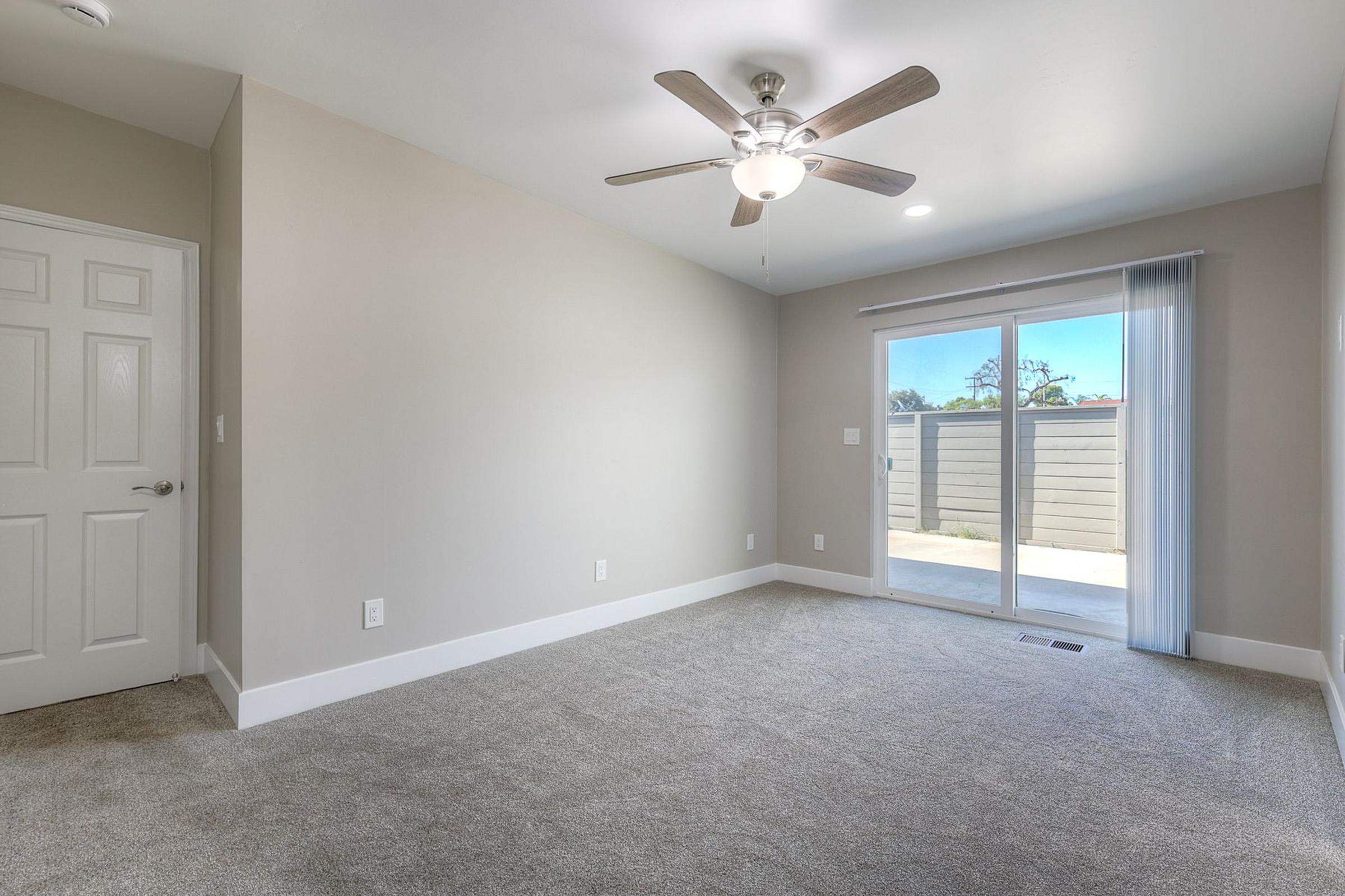A spacious, empty room with light gray walls and plush carpet. It features a ceiling fan and a sliding glass door that opens to an outdoor patio area. Natural light floods in, creating a bright and airy atmosphere. The room is unadorned, ready for personal decor.