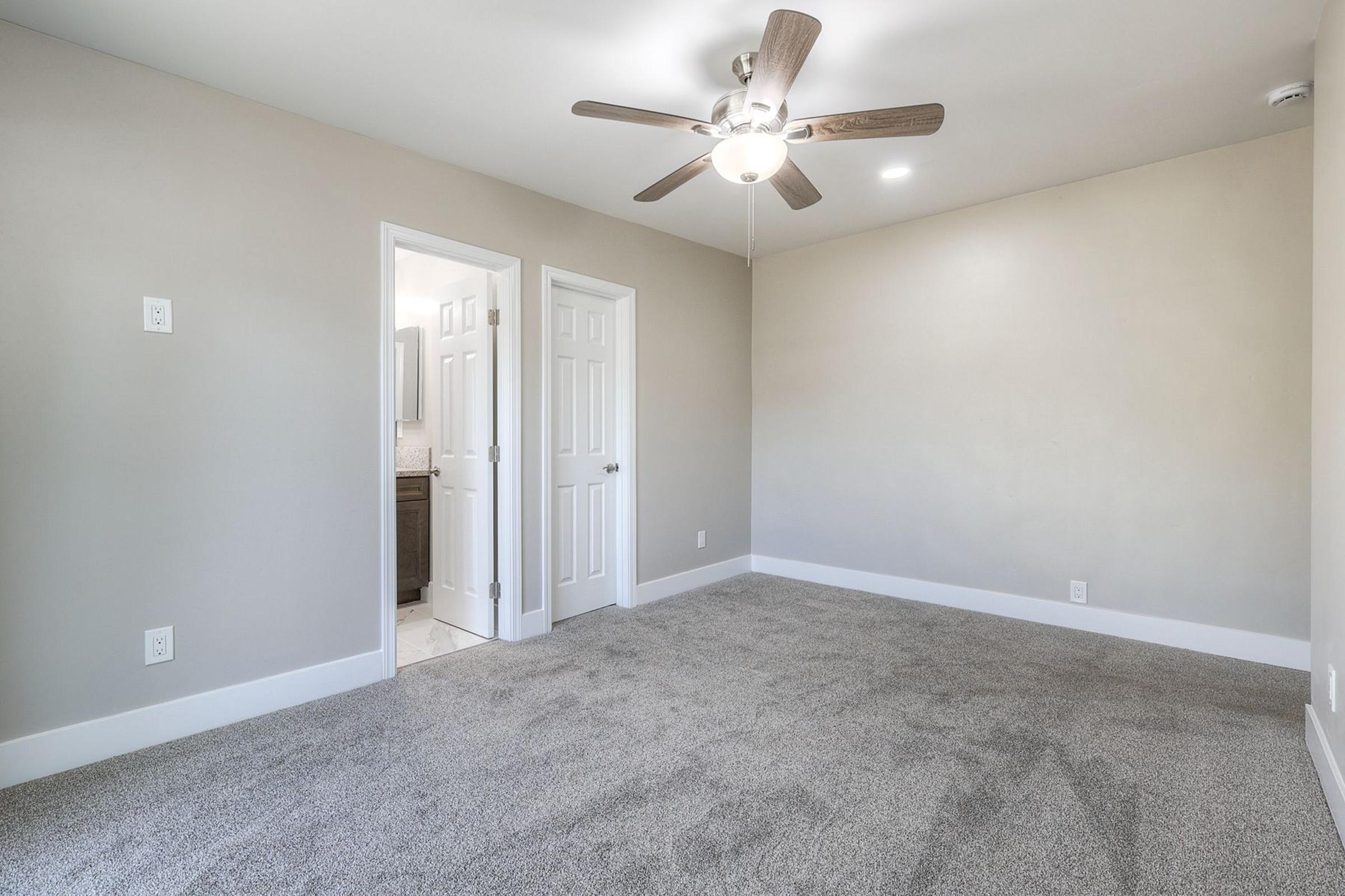 A spacious, carpeted bedroom featuring light gray walls, a ceiling fan with light, and a doorway leading to a bathroom. The room is well-lit with natural light, showcasing a clean and modern design, ideal for relaxation and comfort.
