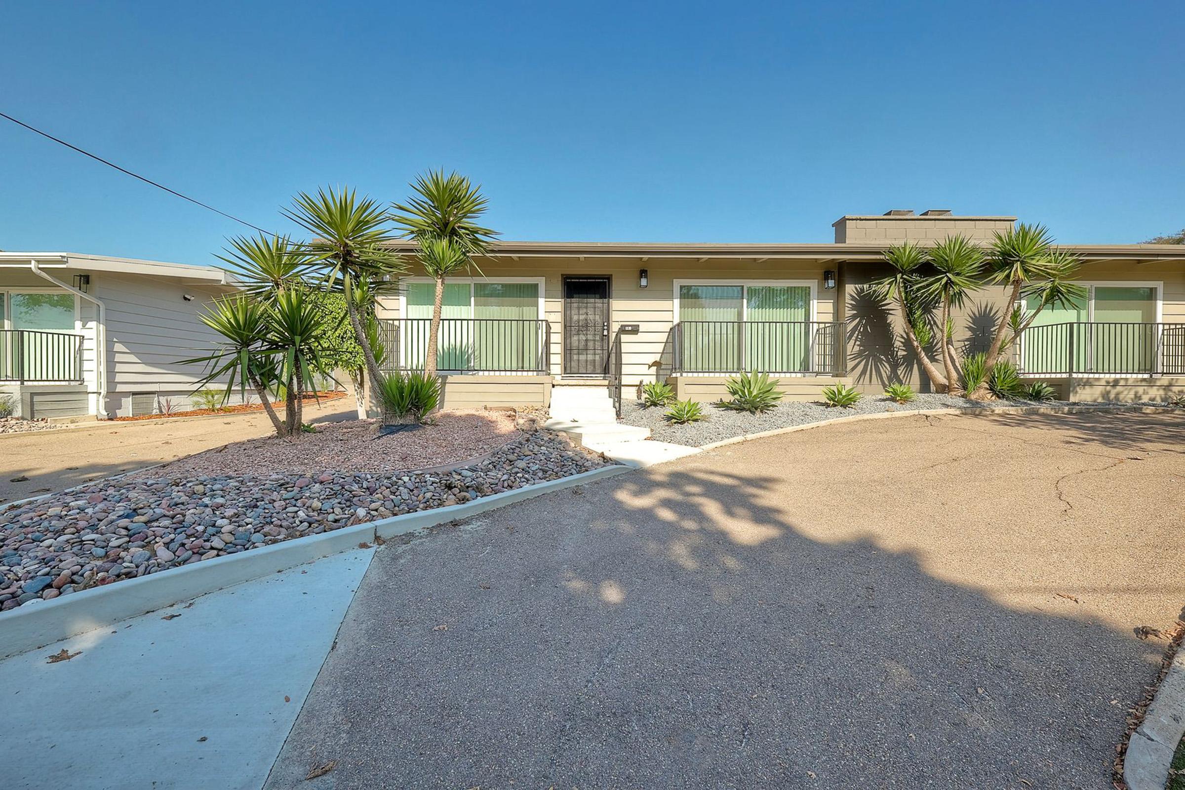 A small, modern house with a landscaped front yard featuring various plants and gravel. The house has a light-colored exterior, large windows, and a set of stairs leading to the entrance. Clear blue skies above enhance the bright and welcoming appearance of the property.