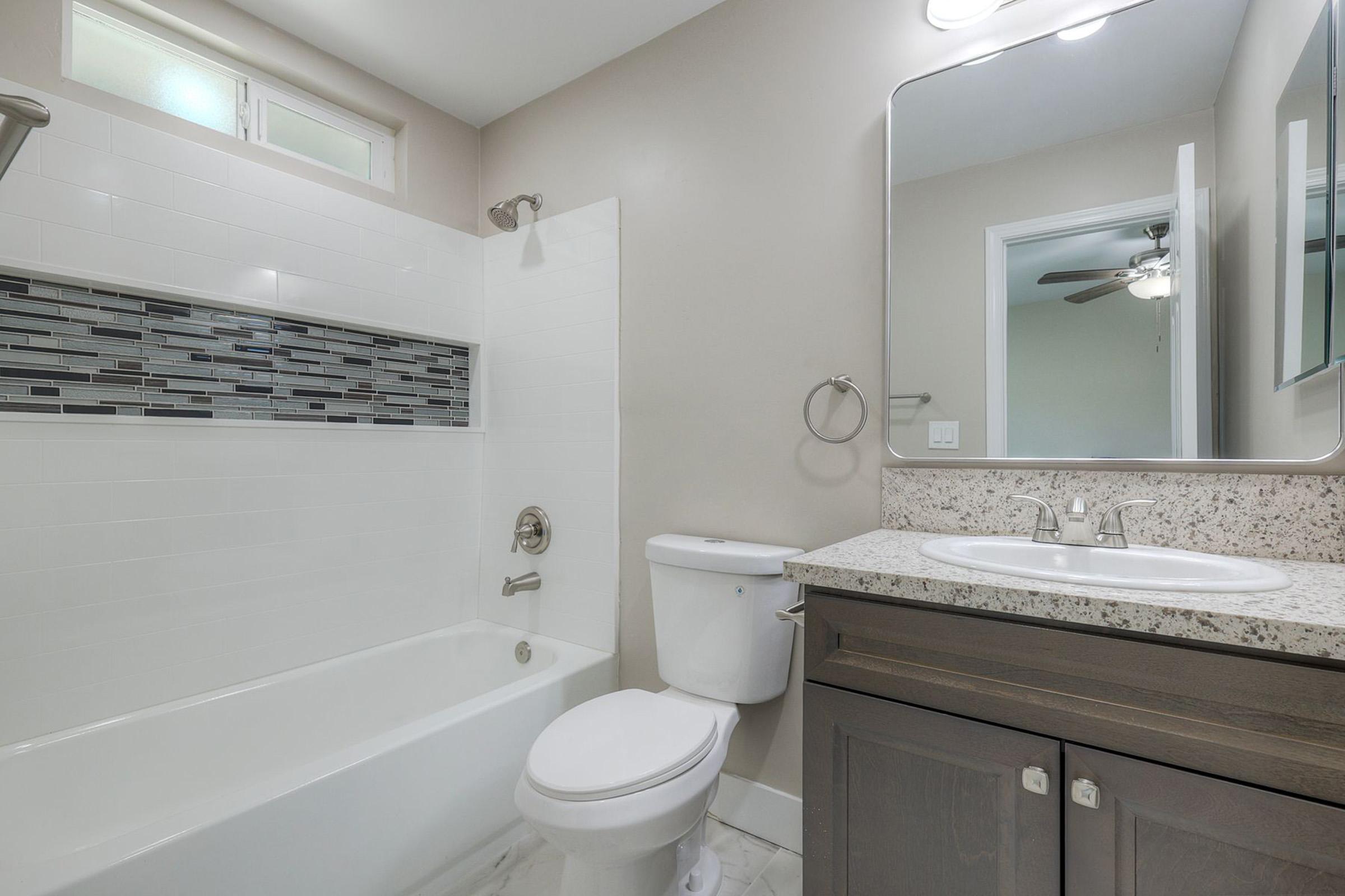 Modern bathroom featuring a bath/shower combo with a decorative tile accent strip, a white toilet, and a granite countertop with dual sinks. The walls are painted a soft color, and a mirror is mounted above the sinks. There is good natural and artificial lighting, enhancing the space's overall brightness.