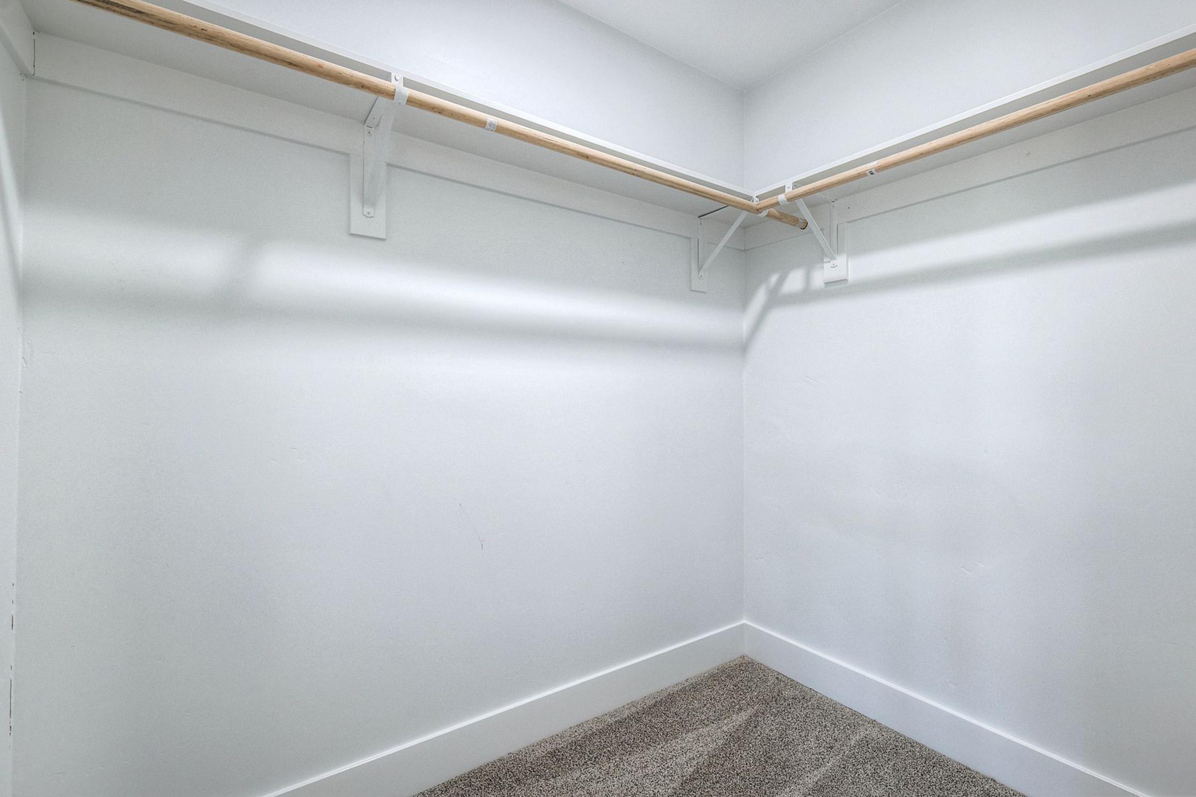 Empty closet with white walls and a light gray carpet. It features a wooden clothing rod mounted on brackets along the wall, providing minimal storage space. The overall look is clean and minimalistic, with no items or furnishings present.