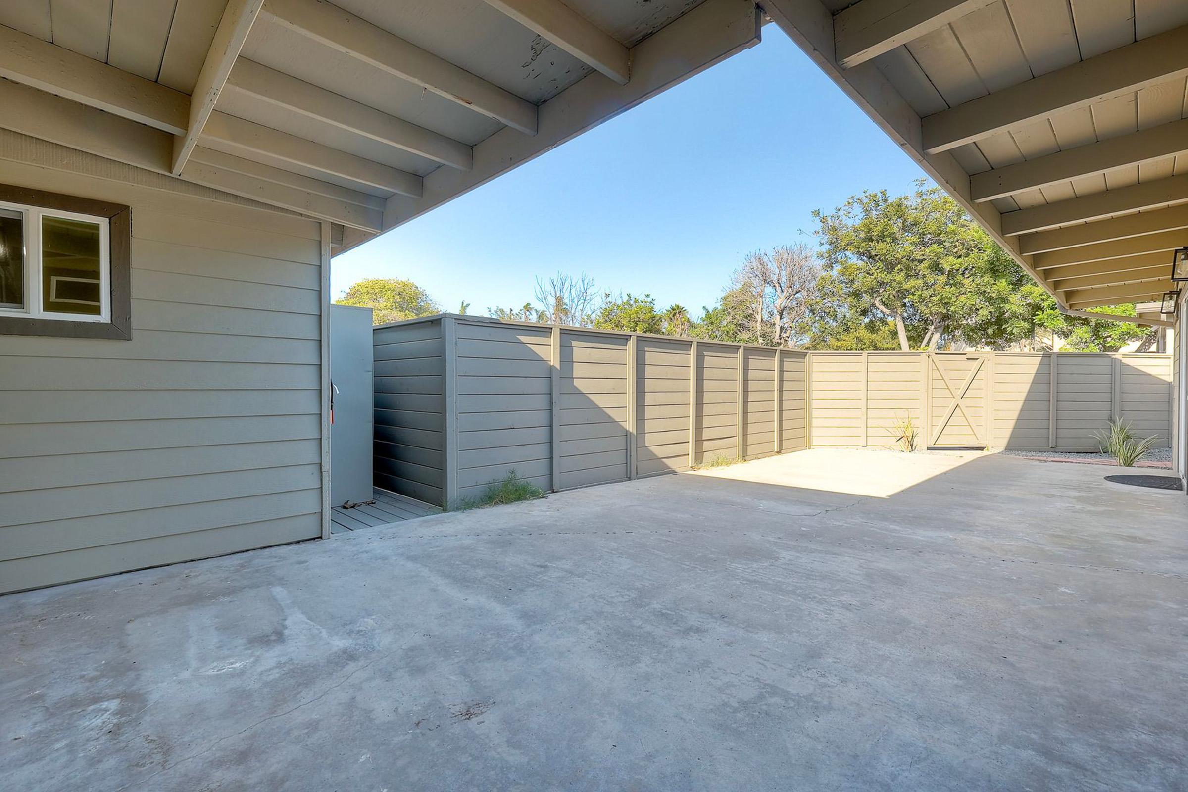 A spacious outdoor area enclosed by wooden fences, featuring a concrete surface and clear blue sky. The scene includes a house on one side and some greenery in the background, creating a serene and open atmosphere.