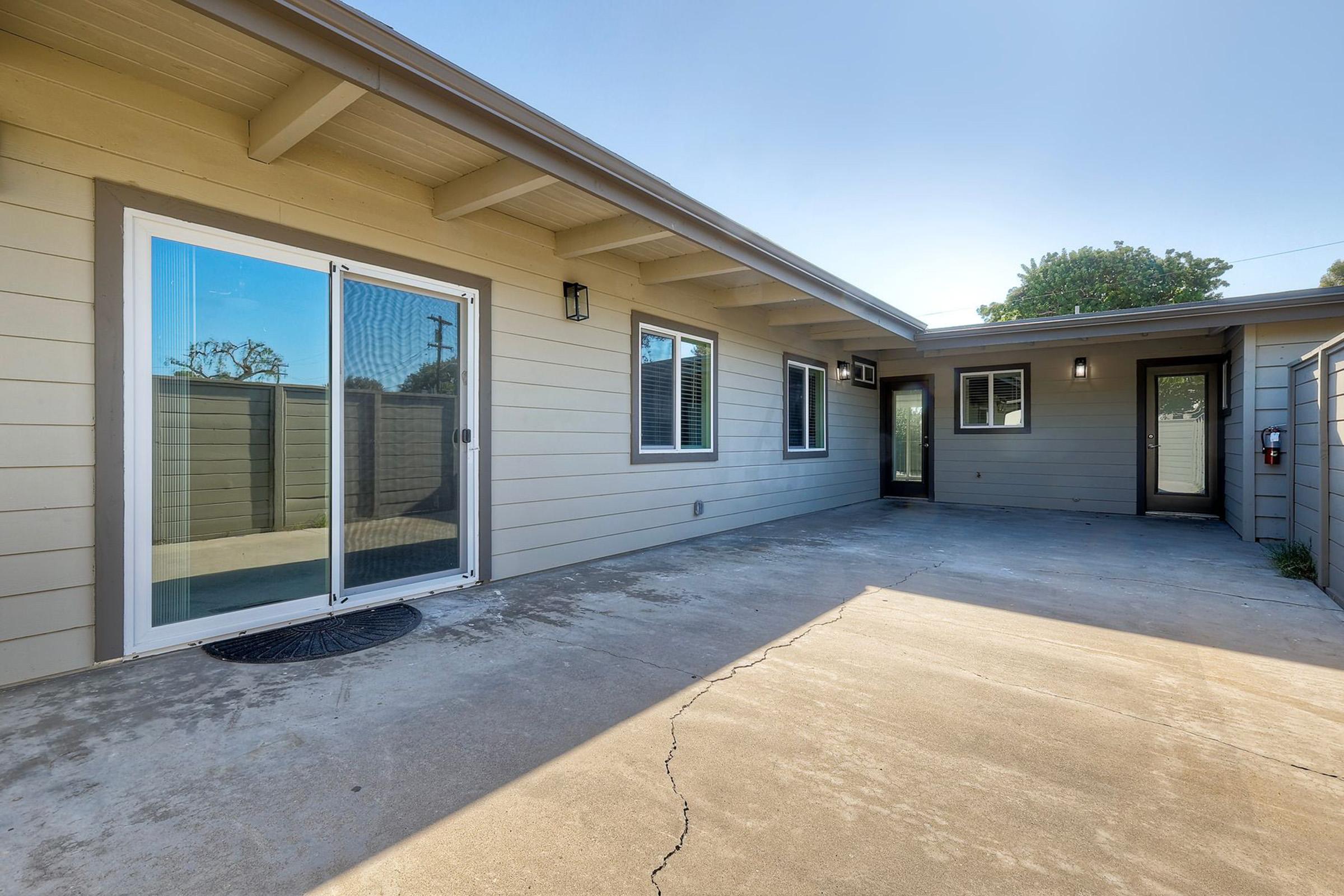 A view of a modern residential patio area featuring gray concrete flooring, sliding glass doors leading to an interior space, and multiple windows. The exterior walls are painted in a light tone, with a neatly maintained fence in the background and clear skies above. The space appears bright and inviting.