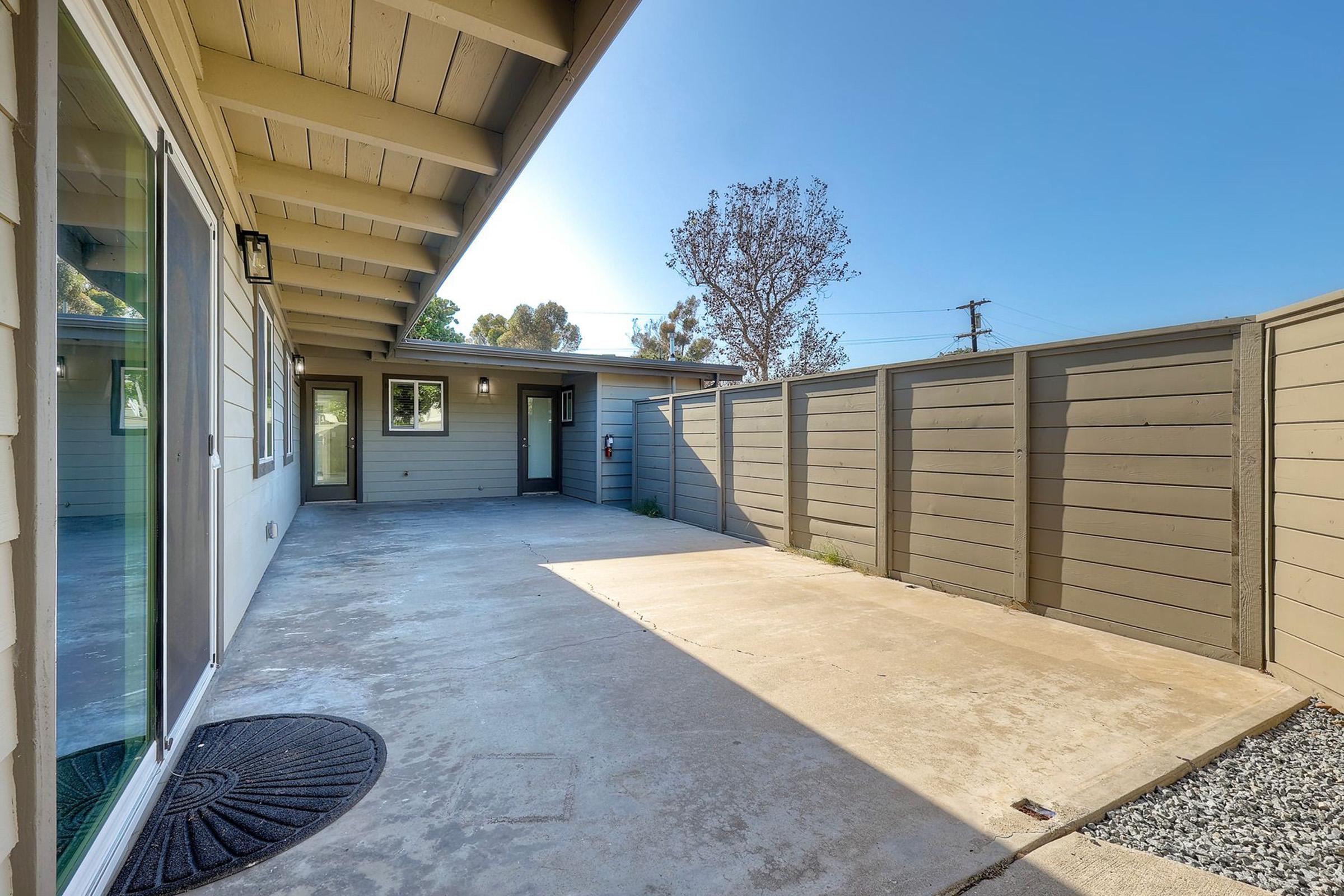 A spacious outdoor area of a modern home featuring a concrete patio, gray fencing, and a tree in the background. The open space is illuminated by sunlight, with a door leading into the house visible. Ideal for outdoor activities or relaxation.