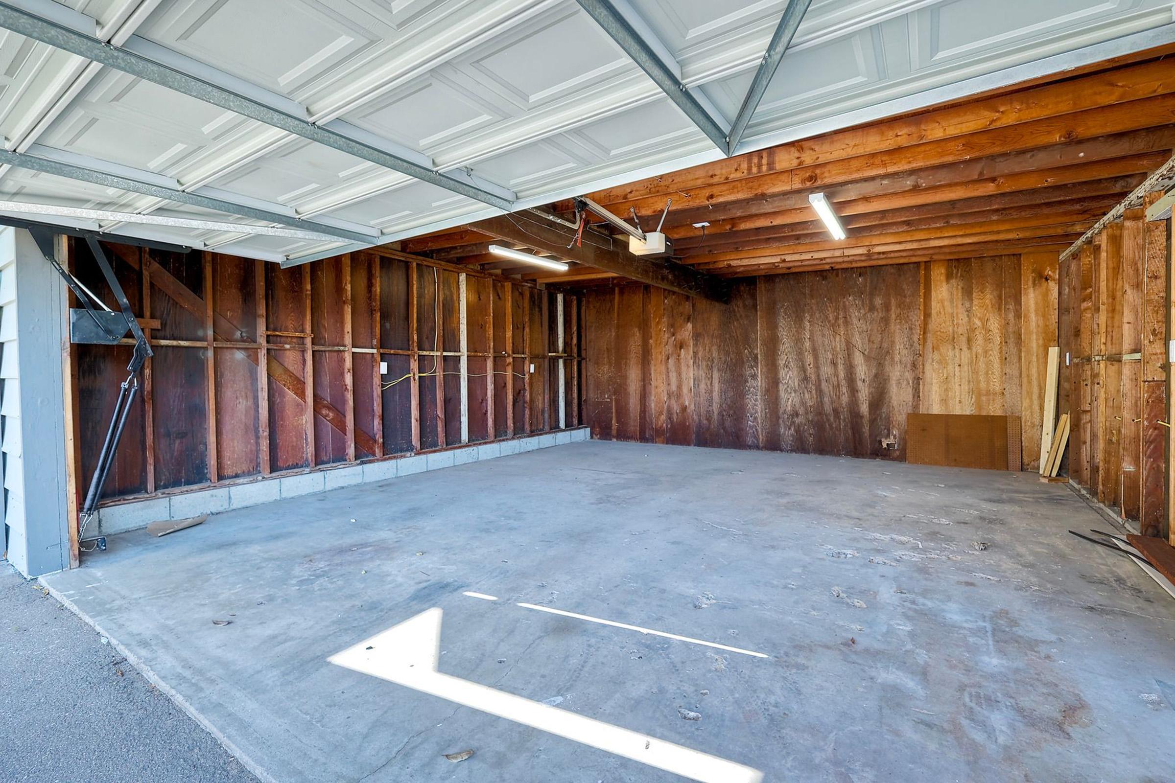 An empty garage with a concrete floor and wooden walls. The garage has a sloped ceiling with fluorescent lights and is devoid of any vehicles or storage items. The door is partially open, allowing natural light to enter.