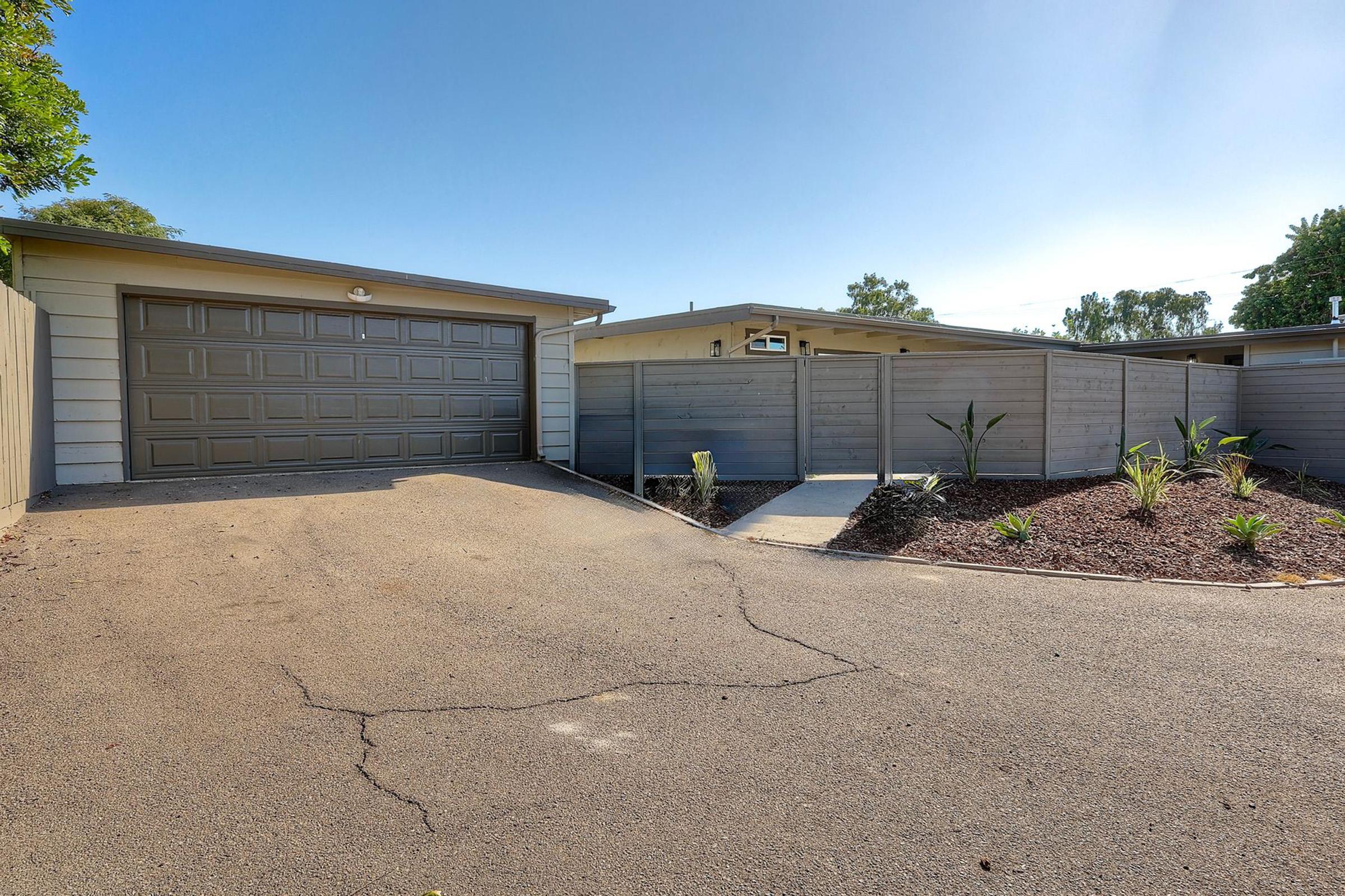 A modern bungalow with a two-car garage, situated on a tree-lined street. The driveway leads up to the garage, and a pathway is visible beside a low fence that separates the yard. Landscaping features include small plants and a gravel area, under a clear blue sky.