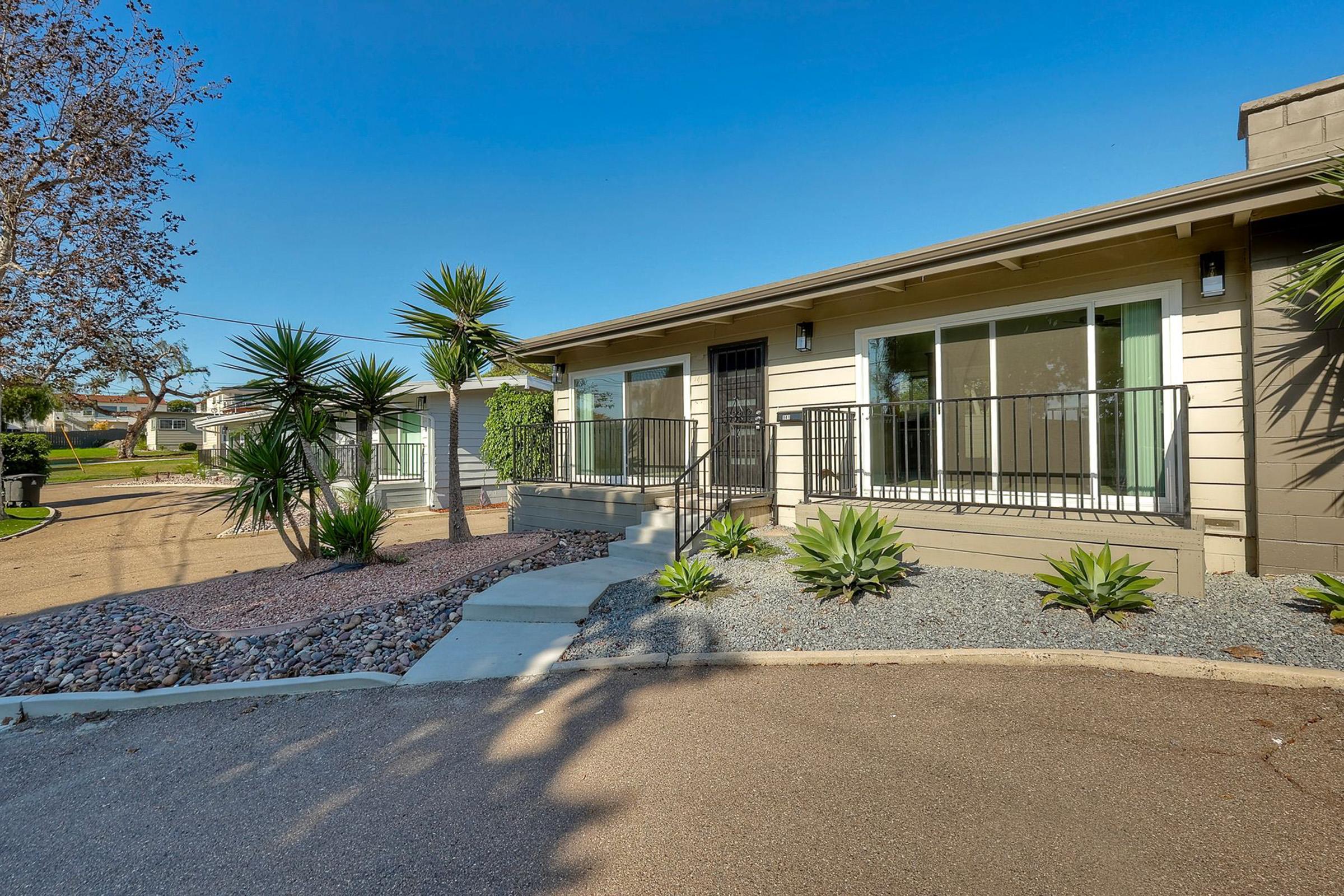 A modern single-story home with a light-colored exterior, featuring large front windows and a black front door. The entrance has a concrete pathway leading up to it, surrounded by landscaped areas with decorative rocks and palm plants. The clear blue sky provides a bright backdrop.