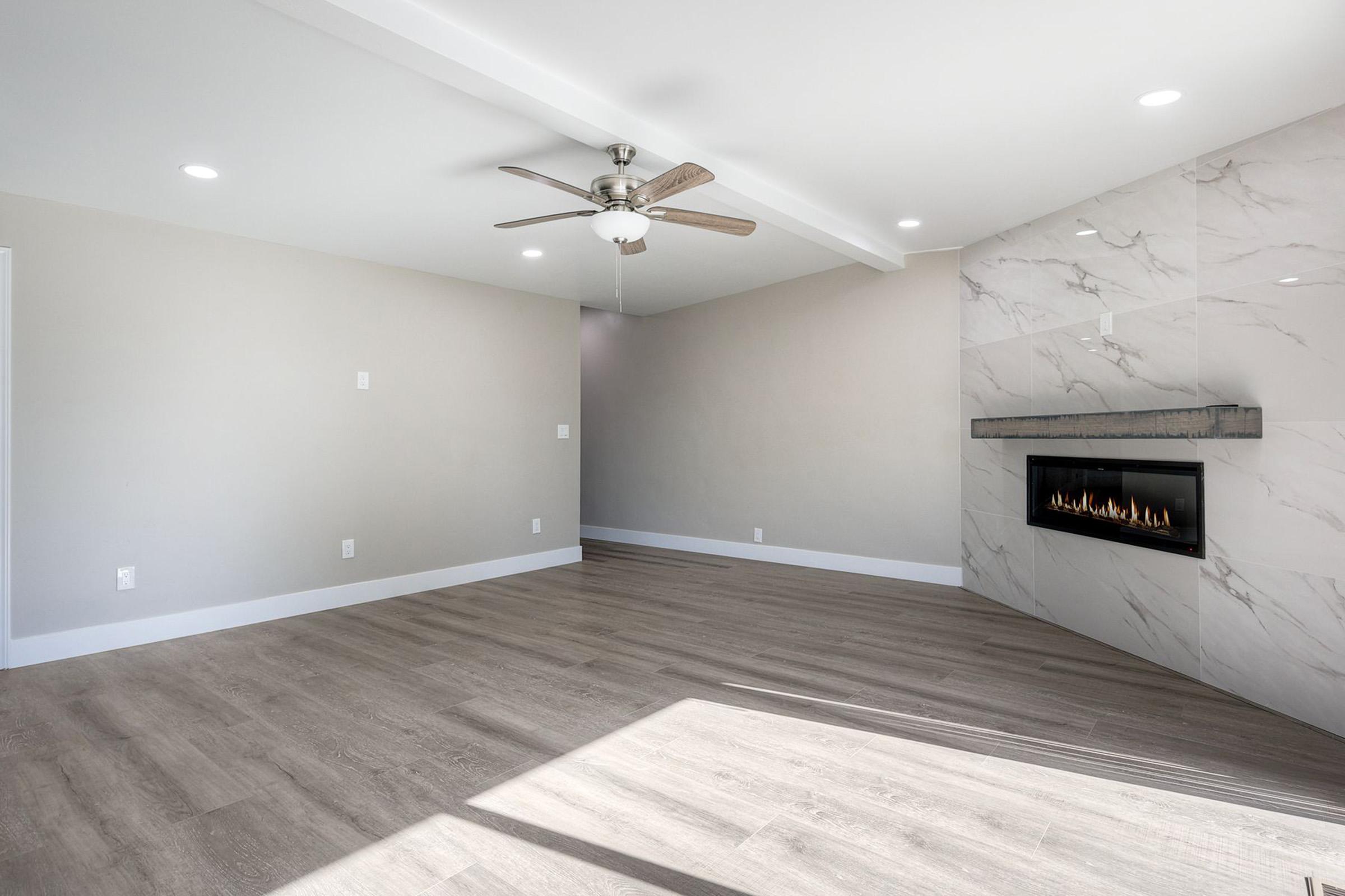 A modern, empty living room featuring light-colored walls, a ceiling fan, and recessed lighting. The floor is hardwood, and a sleek, linear fireplace is set into the wall, complemented by a marble surround. Natural light floods the space, creating a bright and airy atmosphere.