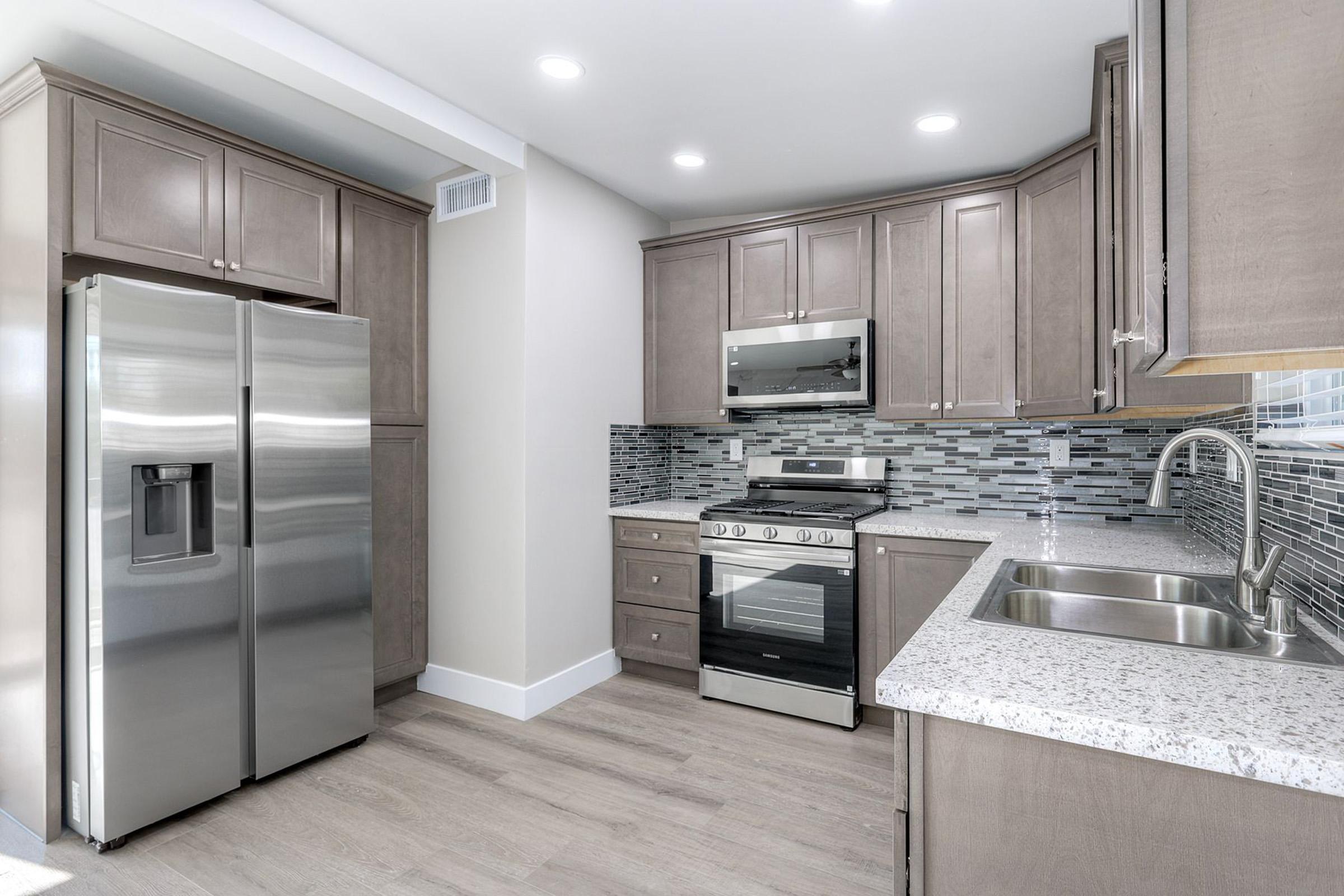 Modern kitchen featuring stainless steel appliances, including a refrigerator and gas range. The cabinets are a light wood finish, complemented by a sleek, speckled granite countertop. A stylish backsplash made of mosaic tiles adds a contemporary touch. The space is well-lit with recessed lighting.