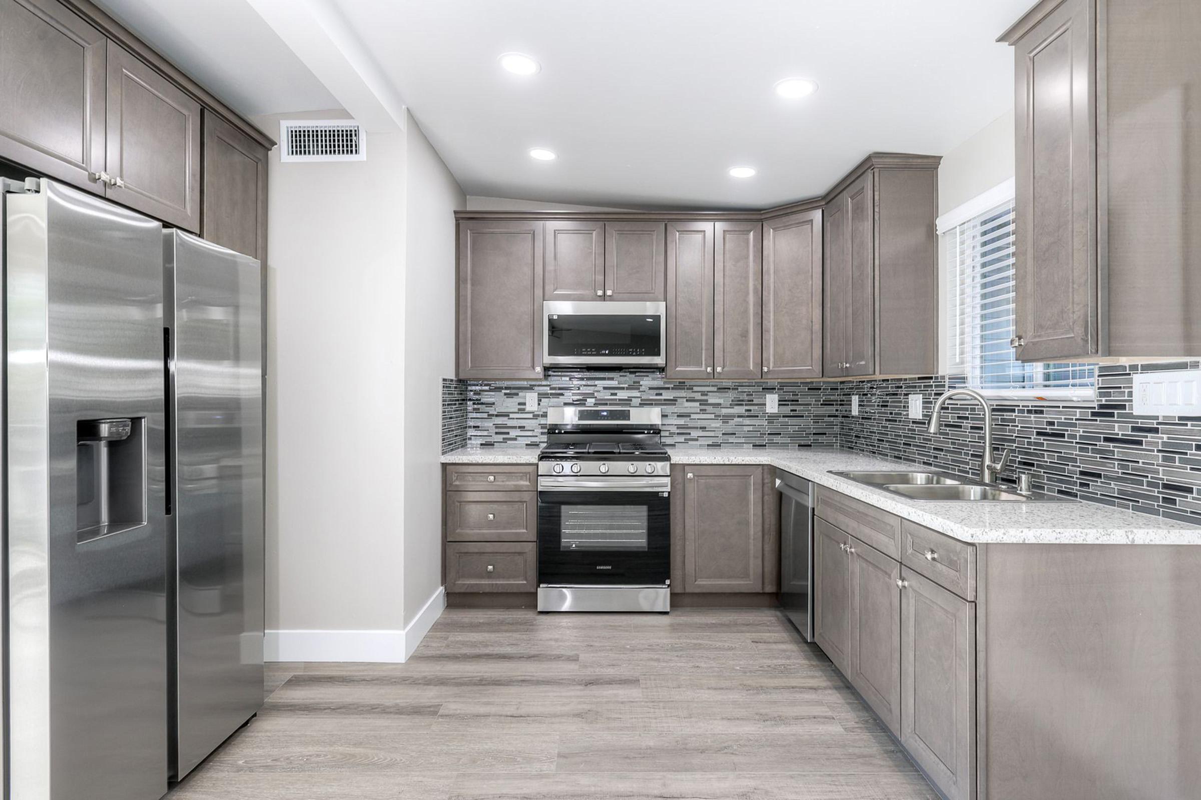 Modern kitchen featuring stainless steel appliances, including a refrigerator and a gas stove. The cabinetry is a warm gray tone, complemented by a mosaic tile backsplash. A spacious countertop provides ample workspace, and natural light streams through a nearby window, enhancing the sleek design.