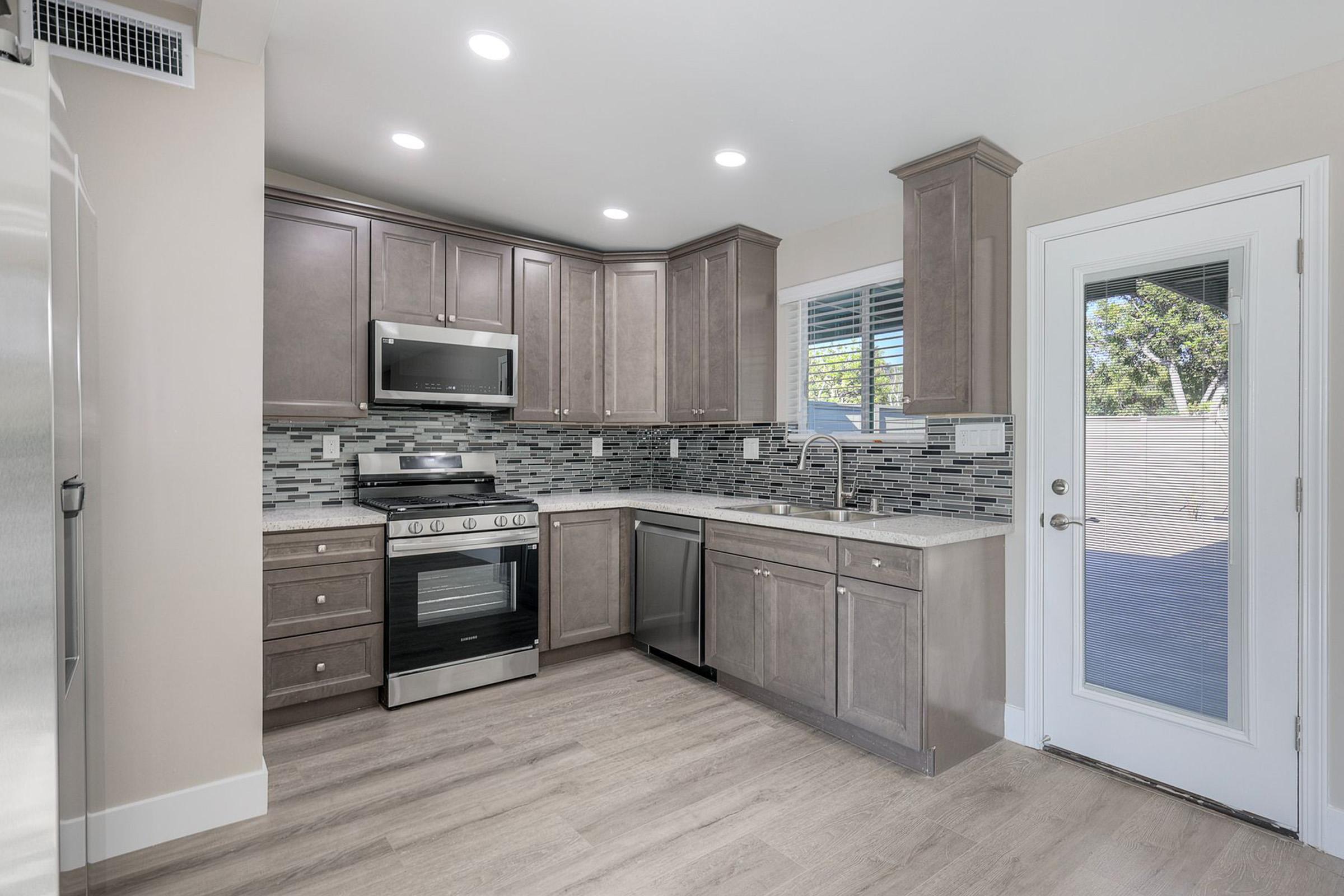 Modern kitchen featuring gray cabinets, stainless steel appliances, and a mosaic backsplash. The design includes a gas stove, built-in microwave, and sleek countertops. Natural light streams in through a window, and there's a door leading outside. The flooring is light-colored wood, enhancing the airy feel.