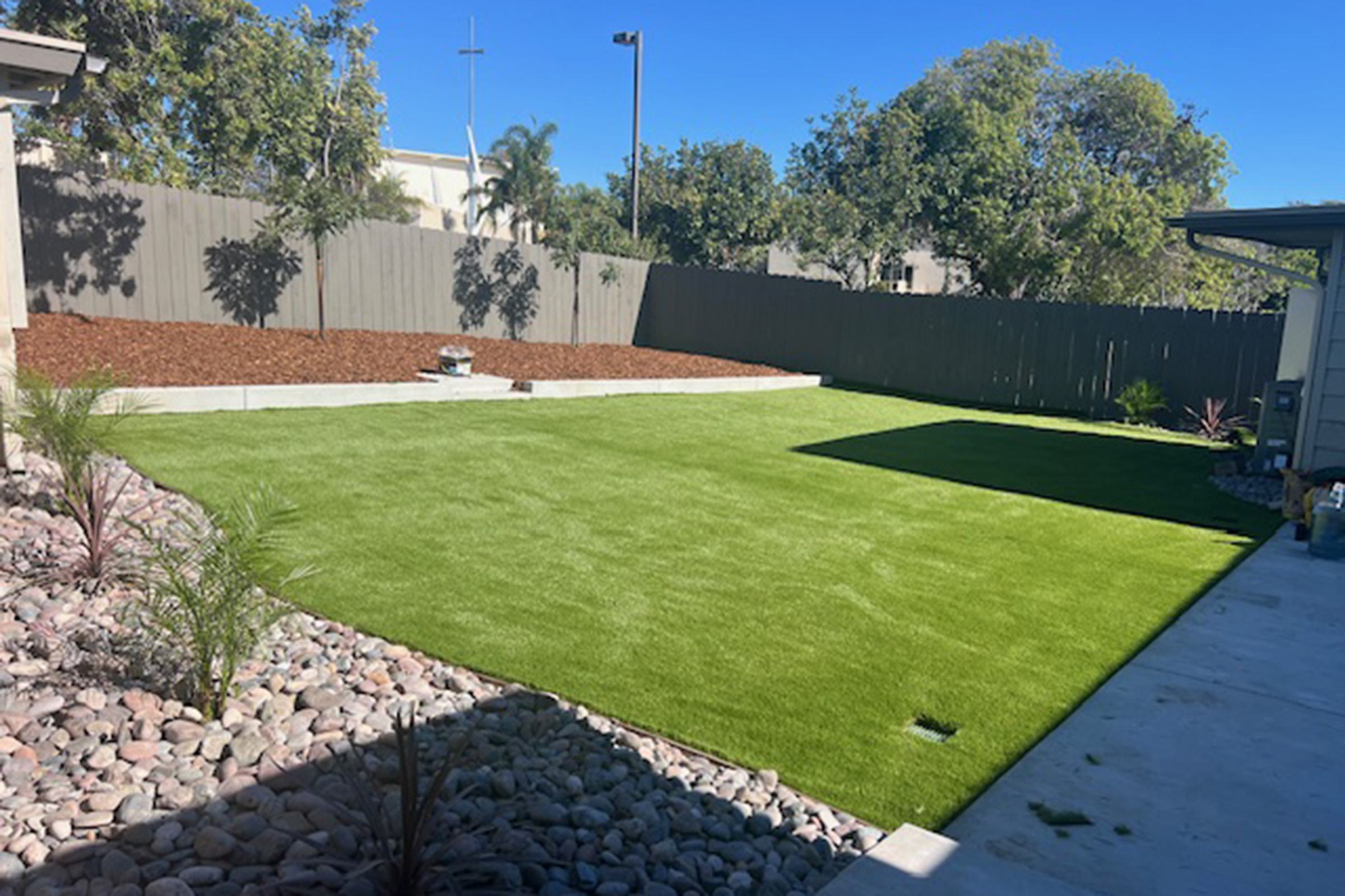 A well-maintained backyard featuring a lush green artificial lawn, bordered by decorative stones. In the background, there are trees and a wooden fence. The area is bright and sunny, creating an inviting outdoor space.