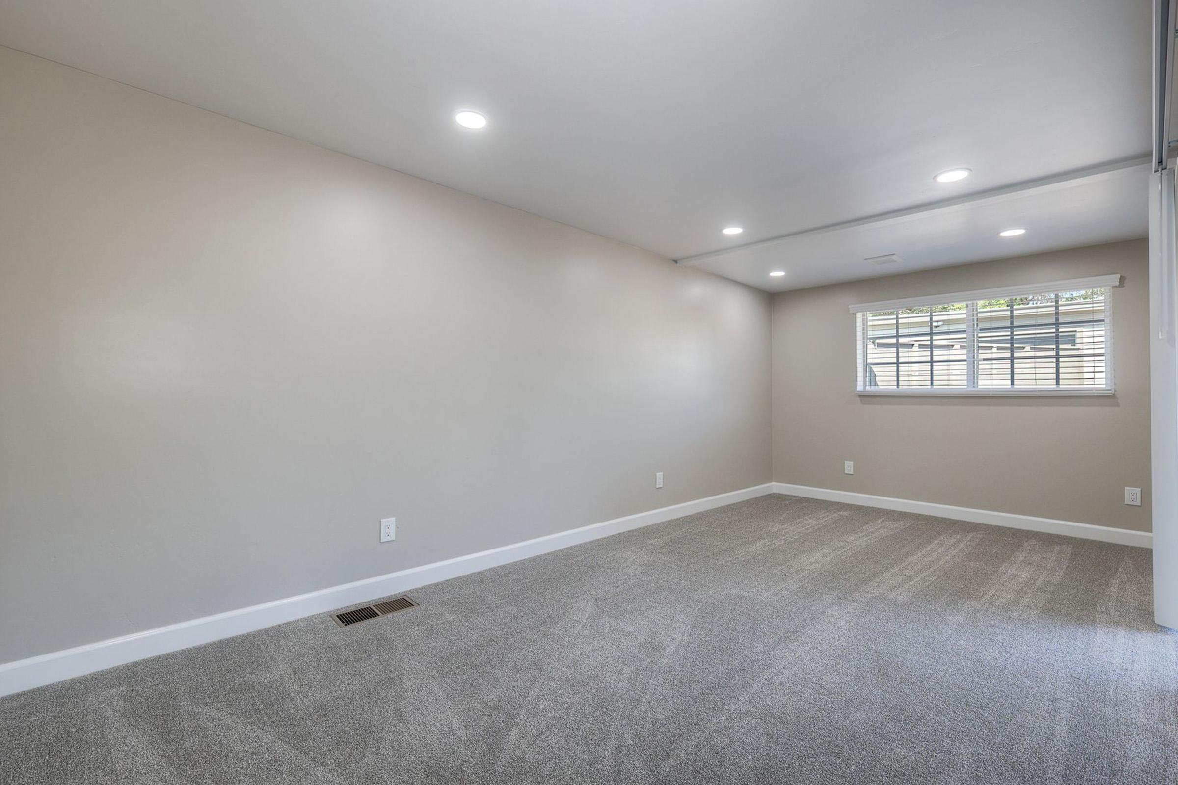 Empty interior of a room with light-colored walls and carpet. There are recessed ceiling lights and a window with white blinds. The space appears well-lit and modern, featuring a clean and minimalistic design.