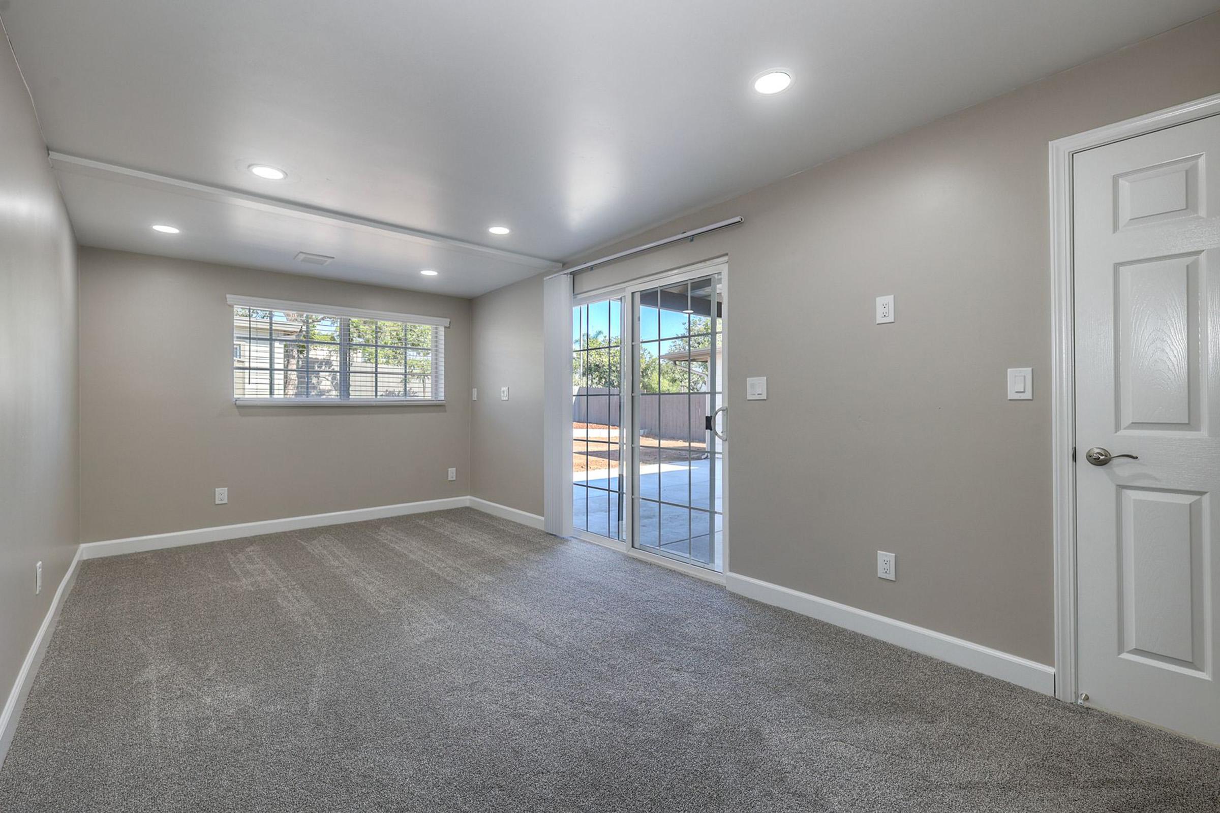 A spacious, empty room with light gray walls and carpet. It features a sliding glass door leading to a patio area, a window with blinds, and recessed lighting on the ceiling. The overall atmosphere is bright and inviting, suitable for various uses.