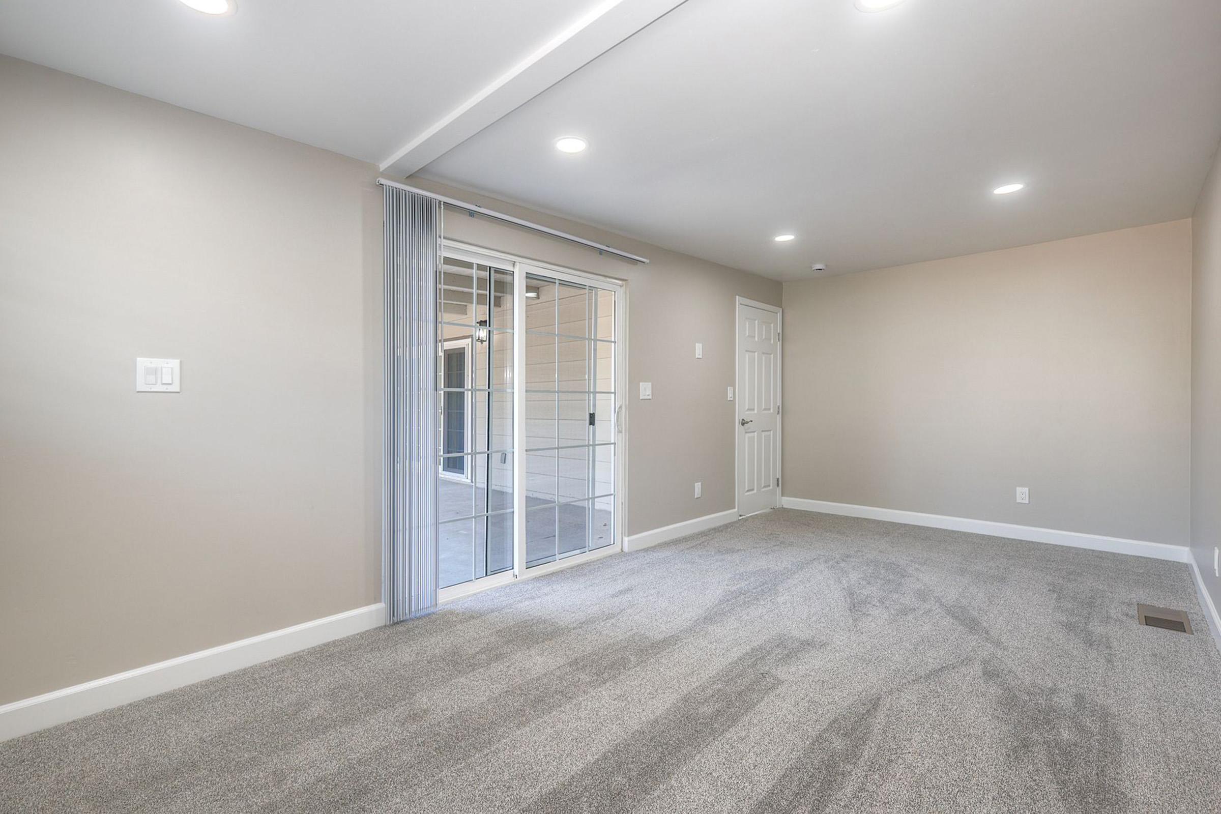 An empty room with beige walls and newly installed gray carpeting. It features large sliding glass doors that open to a patio area, providing natural light. The ceiling has recessed lighting, and there are light switches on the wall. The space appears clean and well-maintained, ideal for various furnishings and decor.
