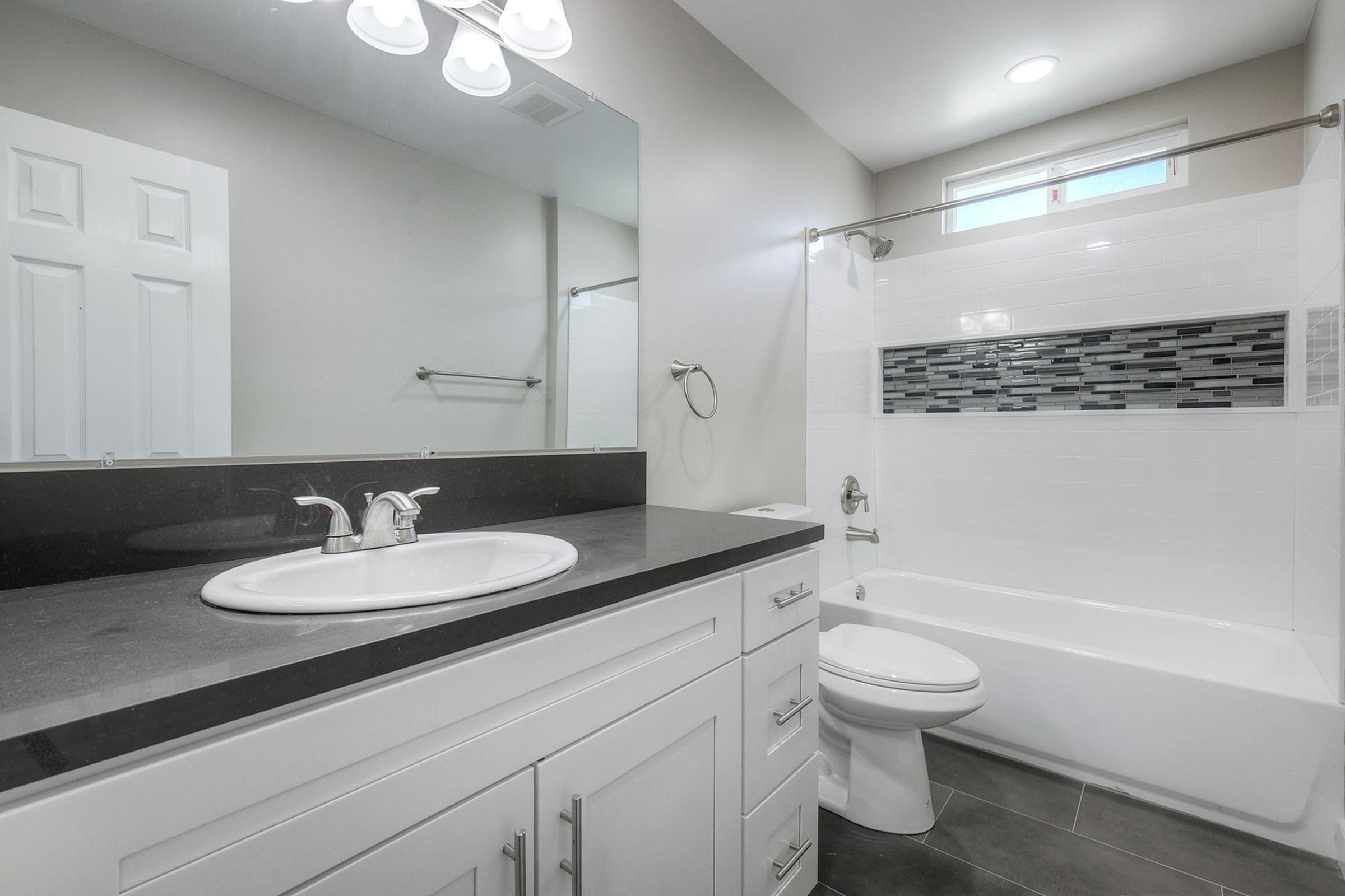 A modern bathroom featuring a white bathtub with a tiled surround, a white sink with a sleek faucet, and a dark countertop. The walls are light-colored, and there is a large mirror above the sink. A small window allows natural light in, and the flooring is dark tiles, creating a contemporary look.