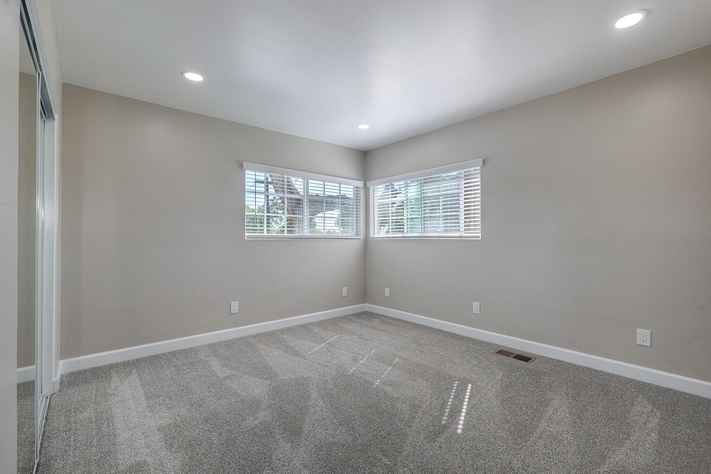 A clean, empty room with light gray carpet and neutral-colored walls. Two windows with white blinds allow natural light to enter, and there is recessed lighting in the ceiling. A wall features a mirrored closet, enhancing the sense of space. The room is ready for personal decoration or furnishings.