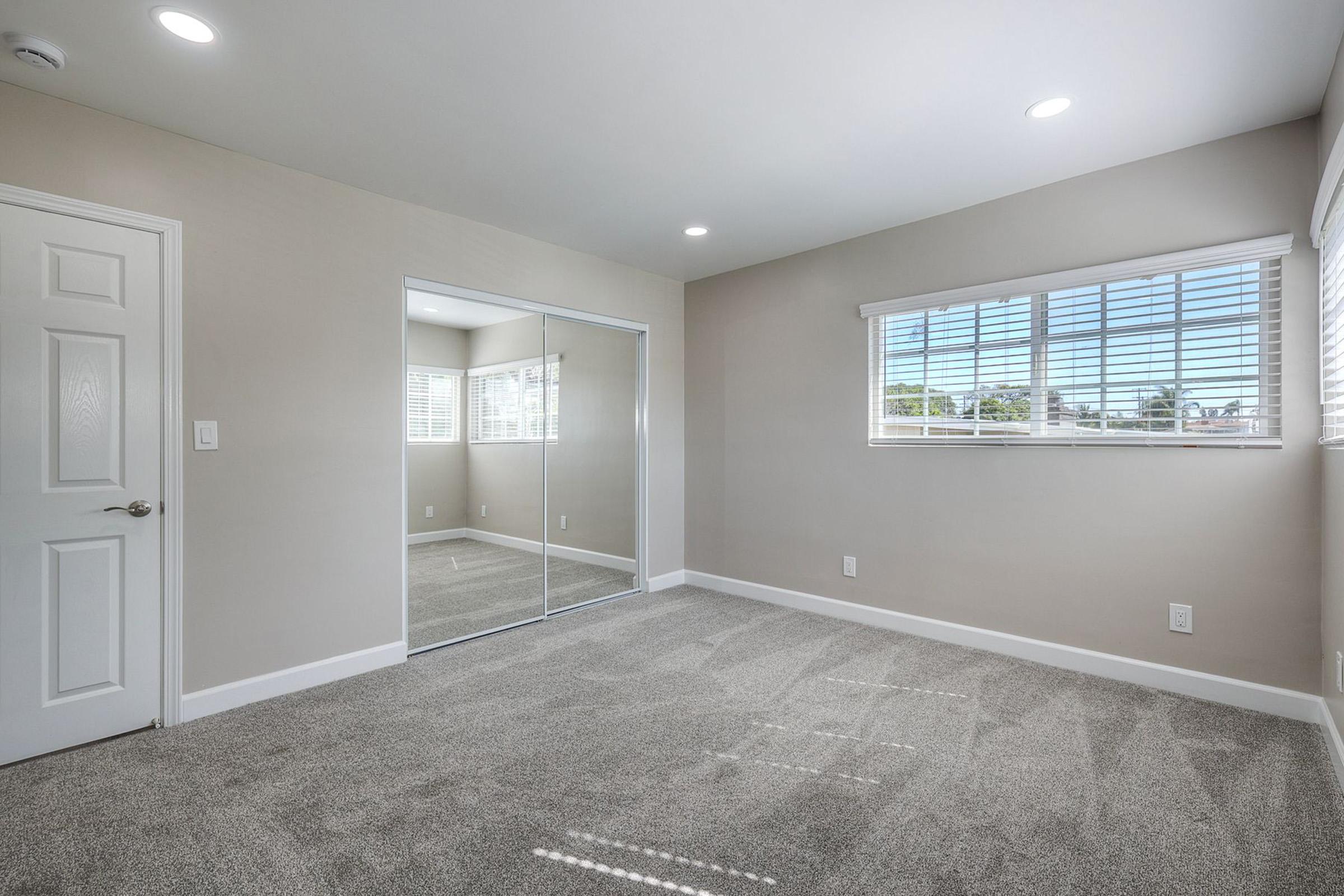 A spacious, empty room with light gray walls and carpet. It features a large window with white blinds, reflecting natural light, and a mirrored closet door. The ceiling has recessed lighting, creating a bright and inviting atmosphere.