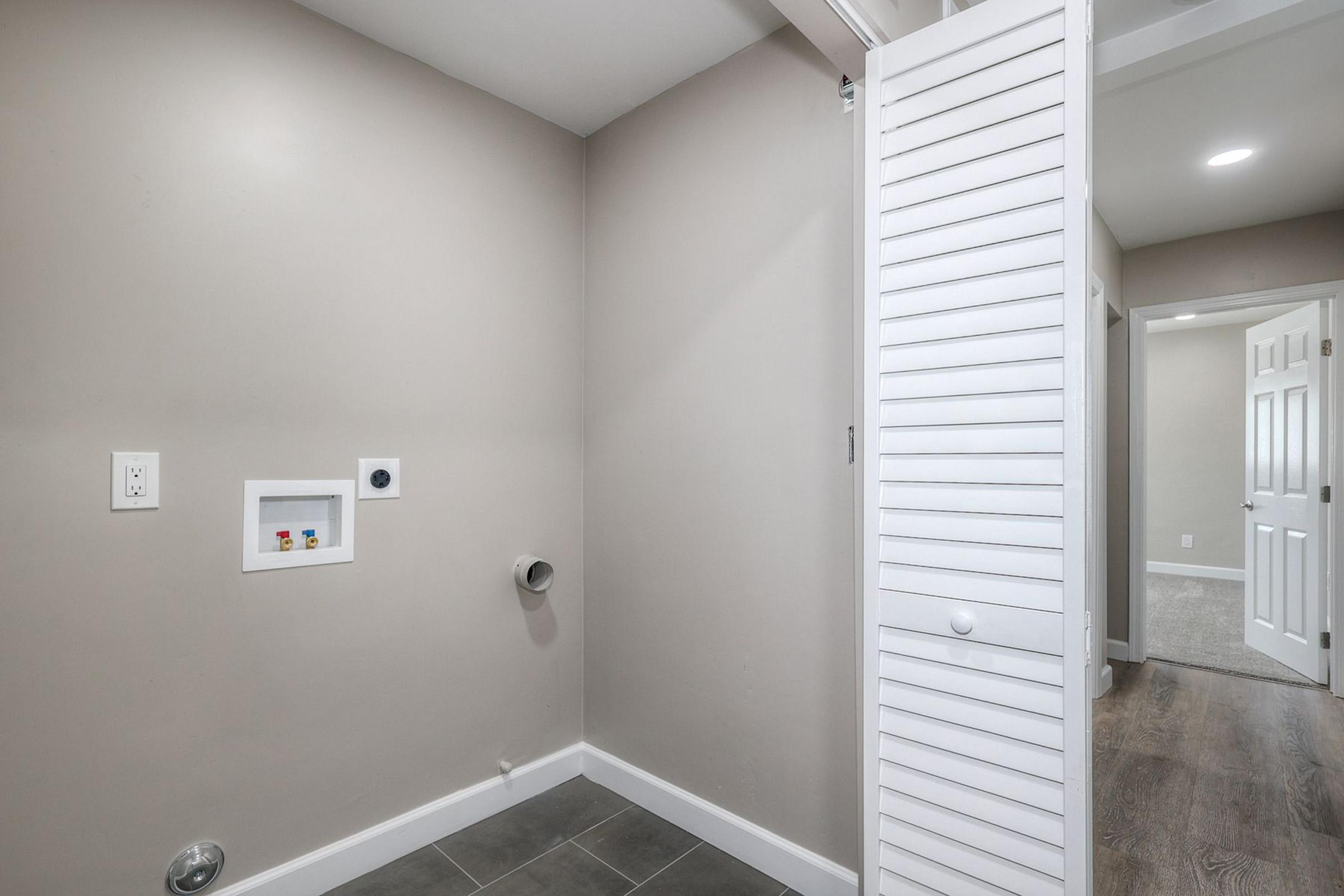 A modern laundry room corner featuring a beige wall, tiled floor, and a white louvered door. There is a small utility panel on the wall and a pipe protruding. A doorway leads to another room, indicating space for laundry appliances and storage. The overall design is clean and well-lit.